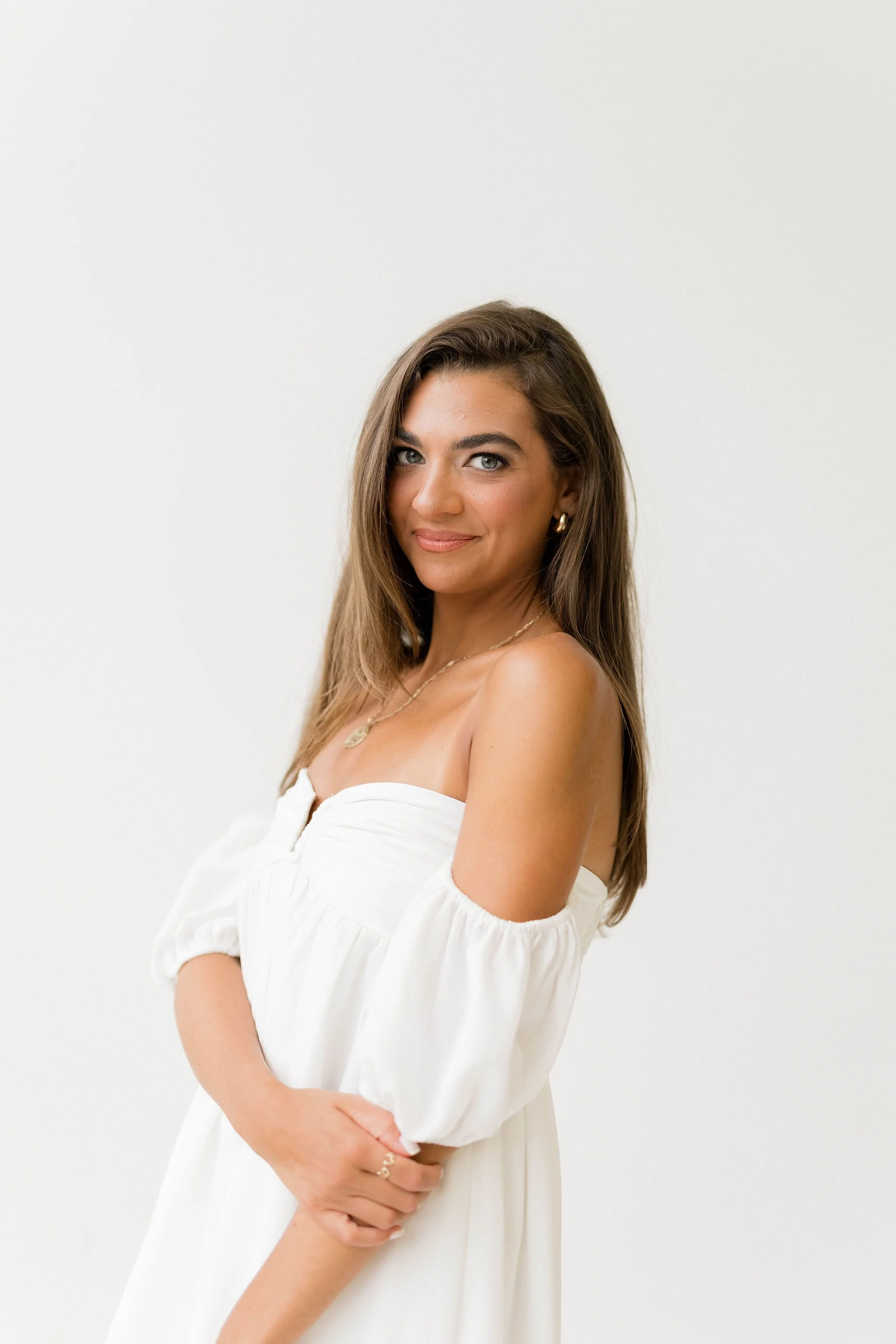A woman with long brown hair, blue eyes, and light skin, wearing a white off-shoulder dress and gold jewelry, smiling softly against a white wall background.