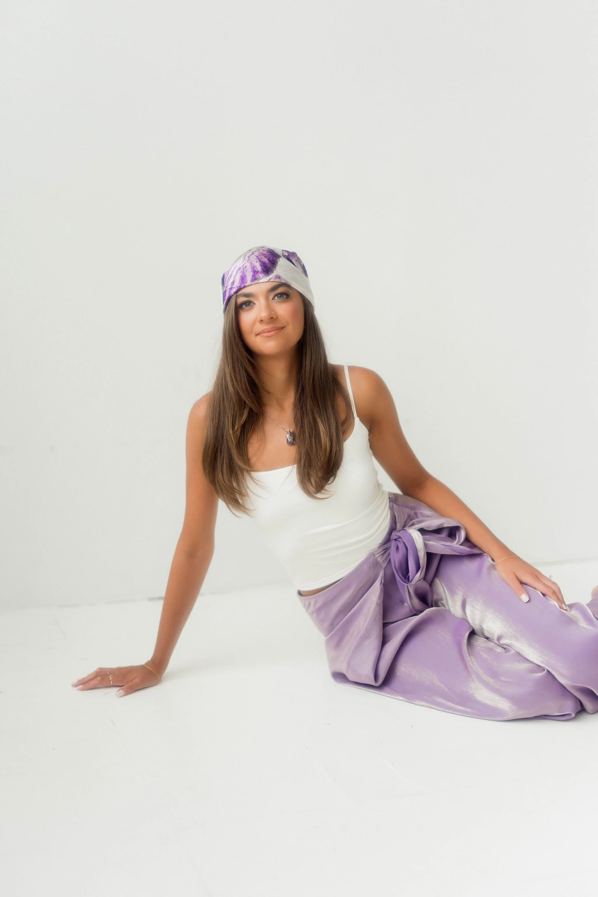 A woman with long brown hair wearing a white tank top, a purple tie-dye bandana, and lavender satin pants, sitting on the floor and smiling against a plain white background.