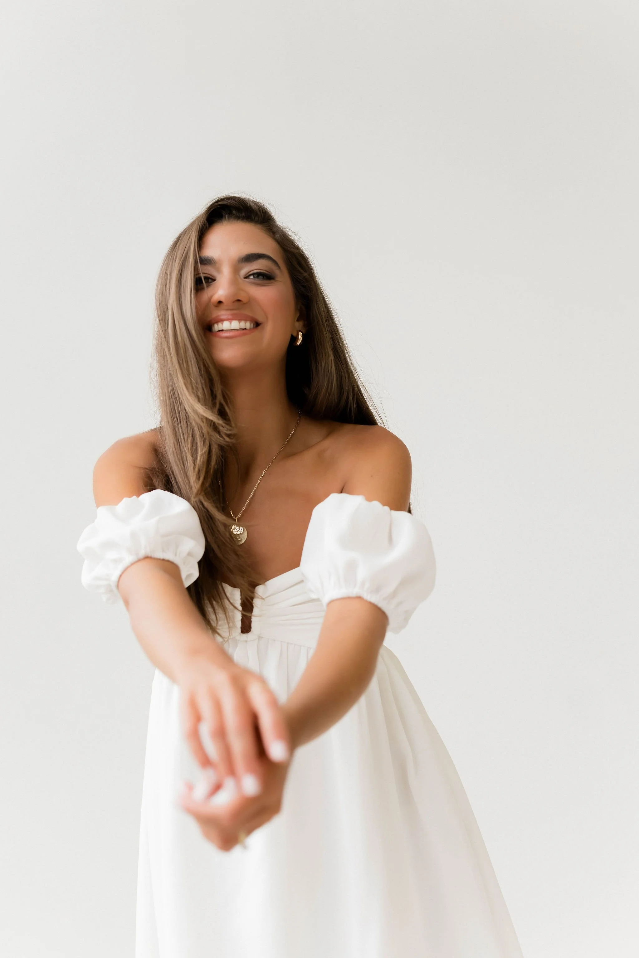 A smiling woman with long brown hair, wearing a white off-the-shoulder dress with puffed sleeves, gold earrings, and a gold necklace, reaching out towards the camera against a plain light-colored background.