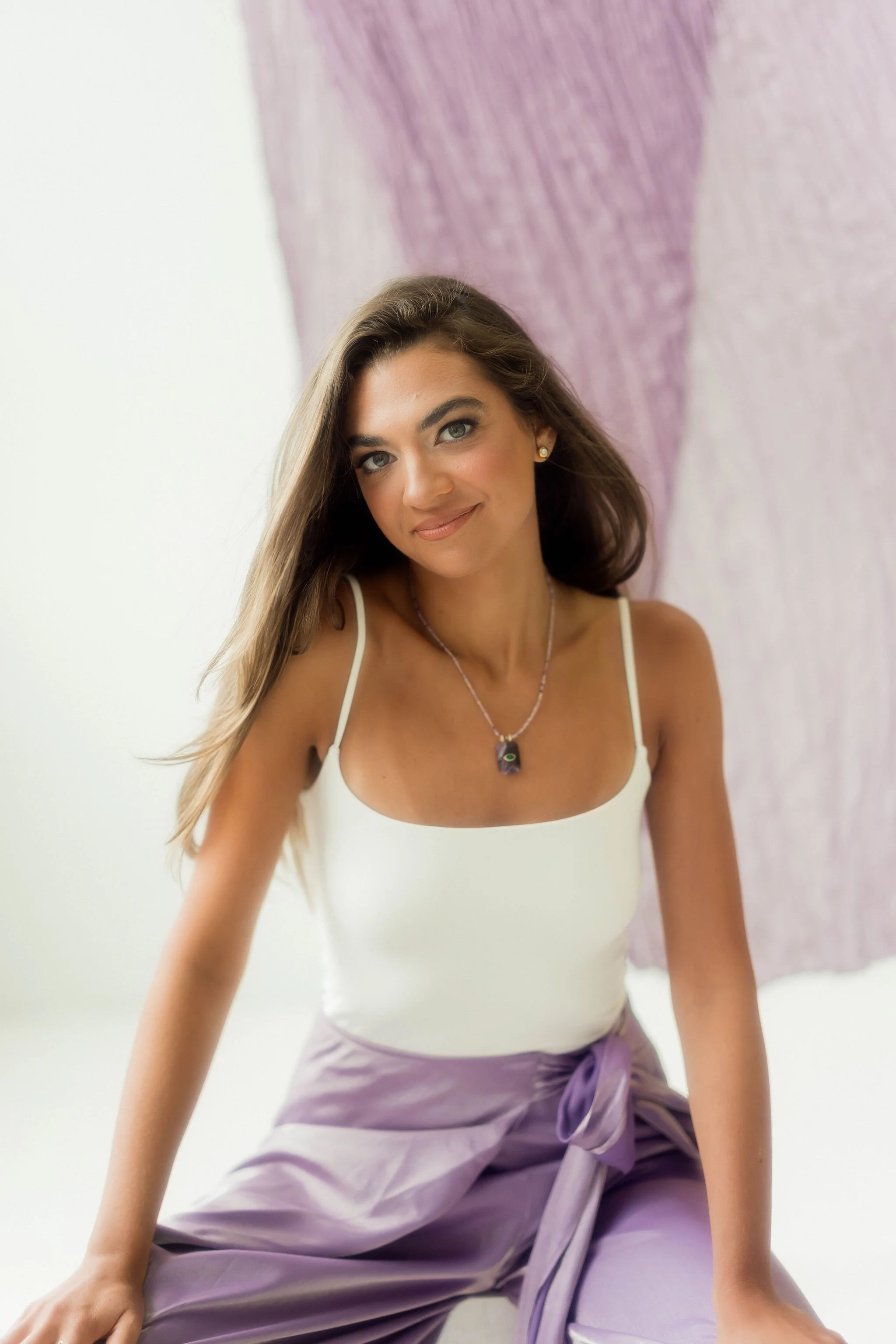 A woman with long brown hair, wearing a white tank top and purple pants with a purple sash, sitting casually in front of a light-colored wall and purple artwork.