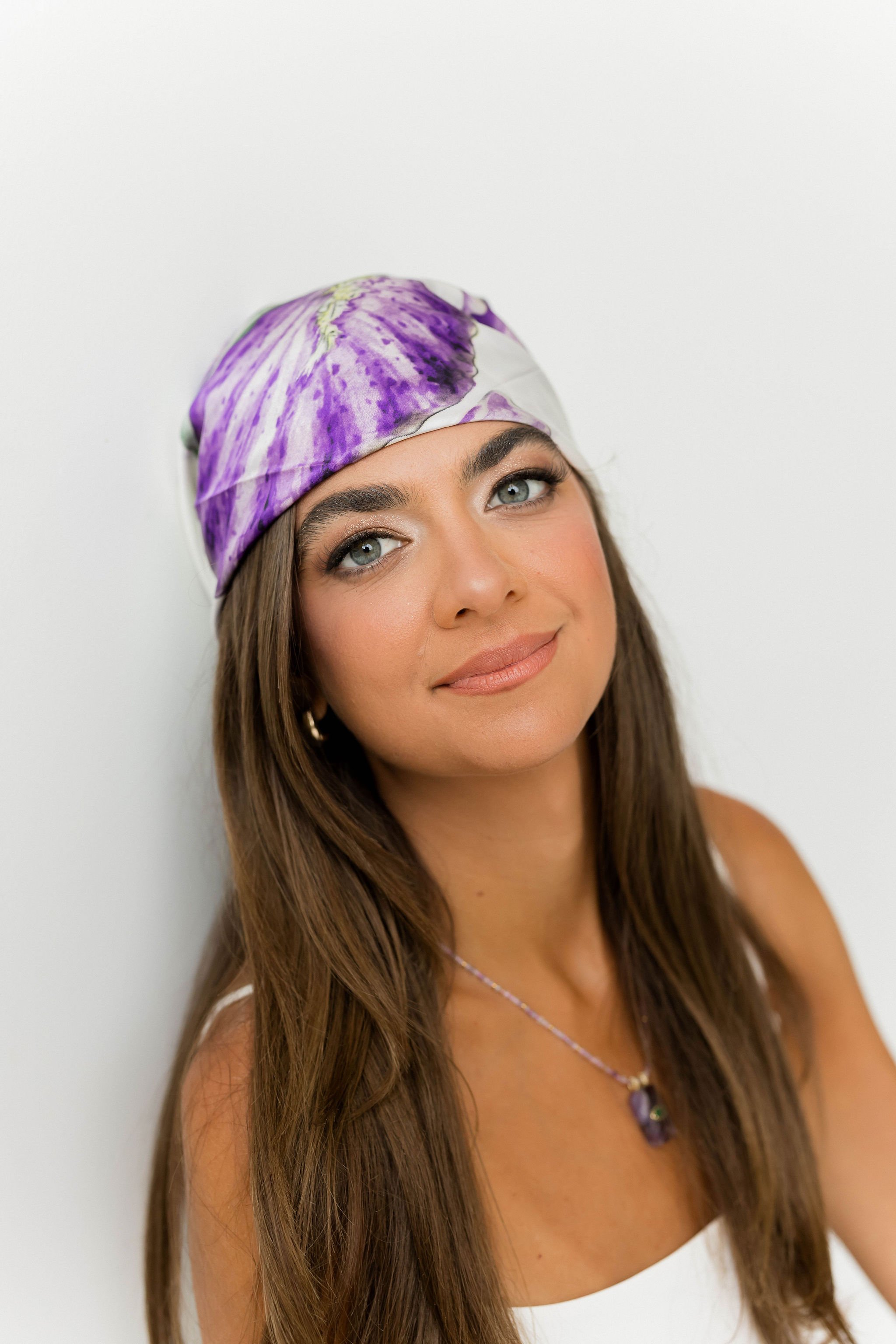 A woman with long brown hair, blue eyes, and a light tan complexion wearing a purple and white headscarf, a white top, and a purple necklace, smiling softly against a plain white background.
