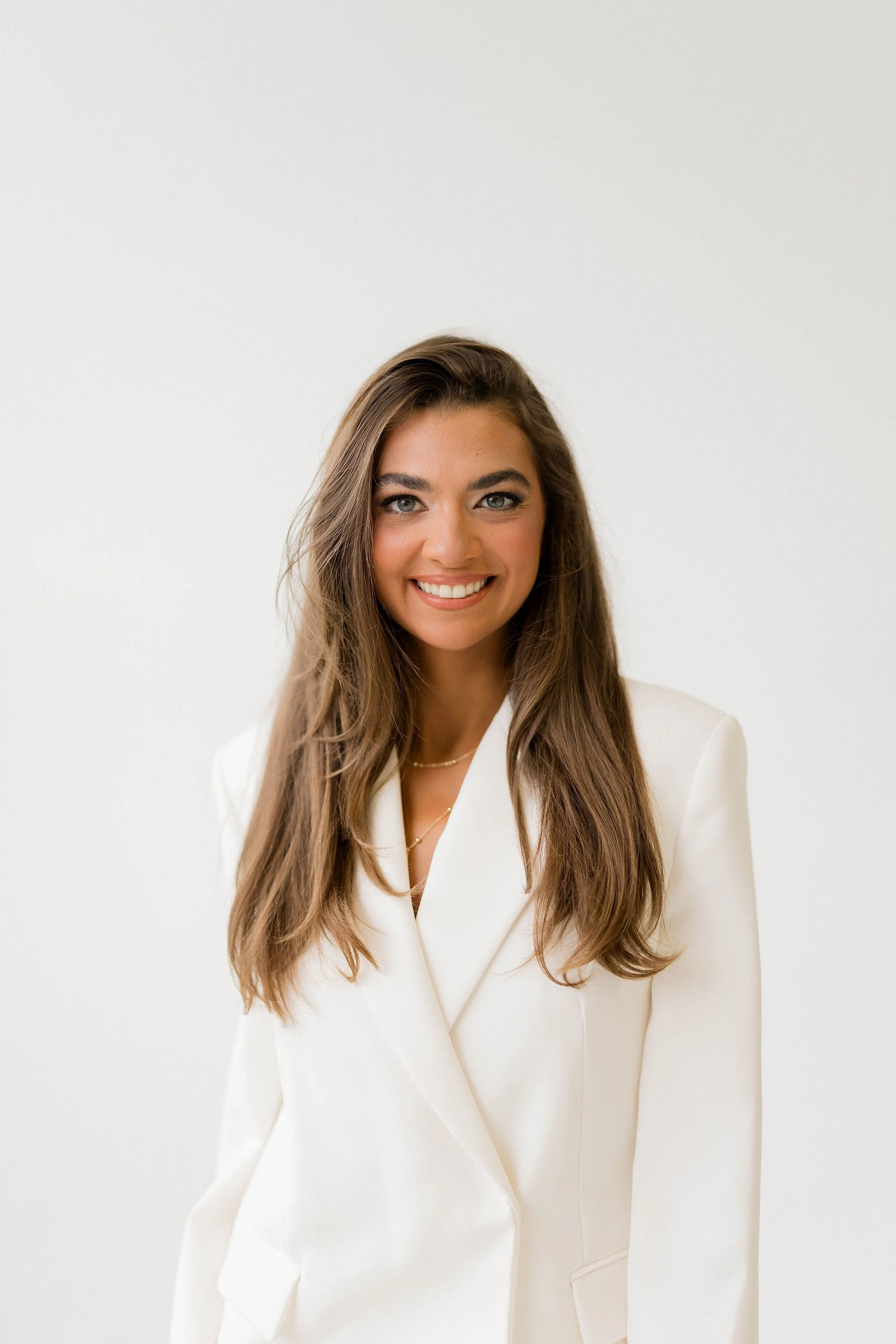 A woman with long brown hair, blue eyes, and a bright smile wearing a white blazer against a plain white background.