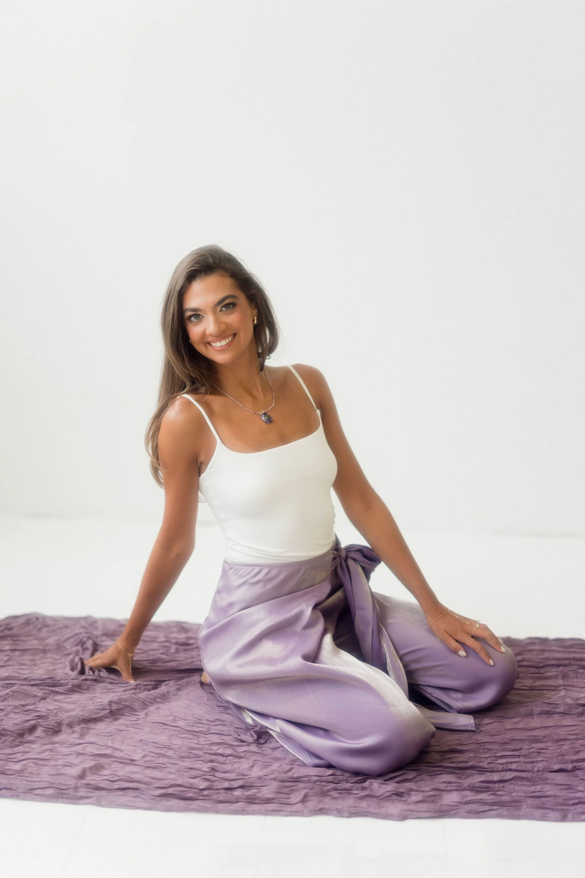 A woman with long brown hair, wearing a white tank top, purple silk pants, and a necklace, sitting on a purple textured blanket against a plain white wall, smiling at the camera.