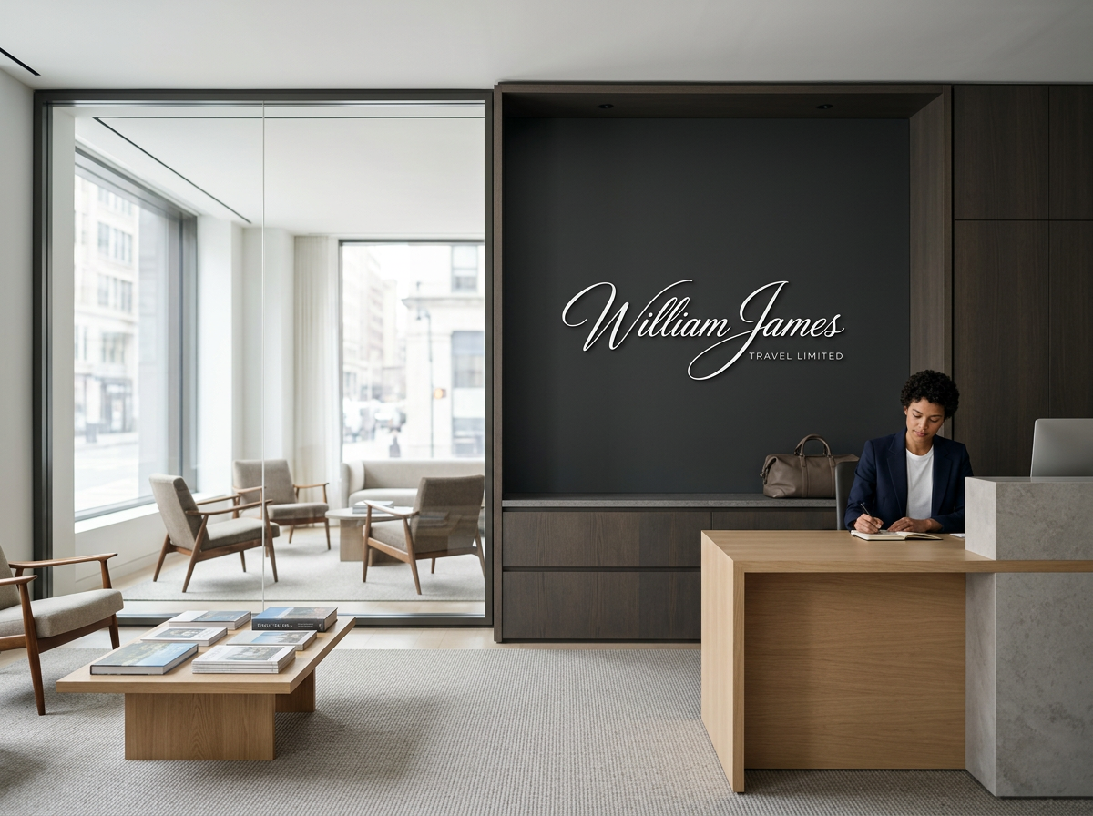 Modern office reception area with a woman sitting at the front desk writing, a dark wall with William James Travel Limited signage, and a sitting area visible through glass doors with chairs and a coffee table.