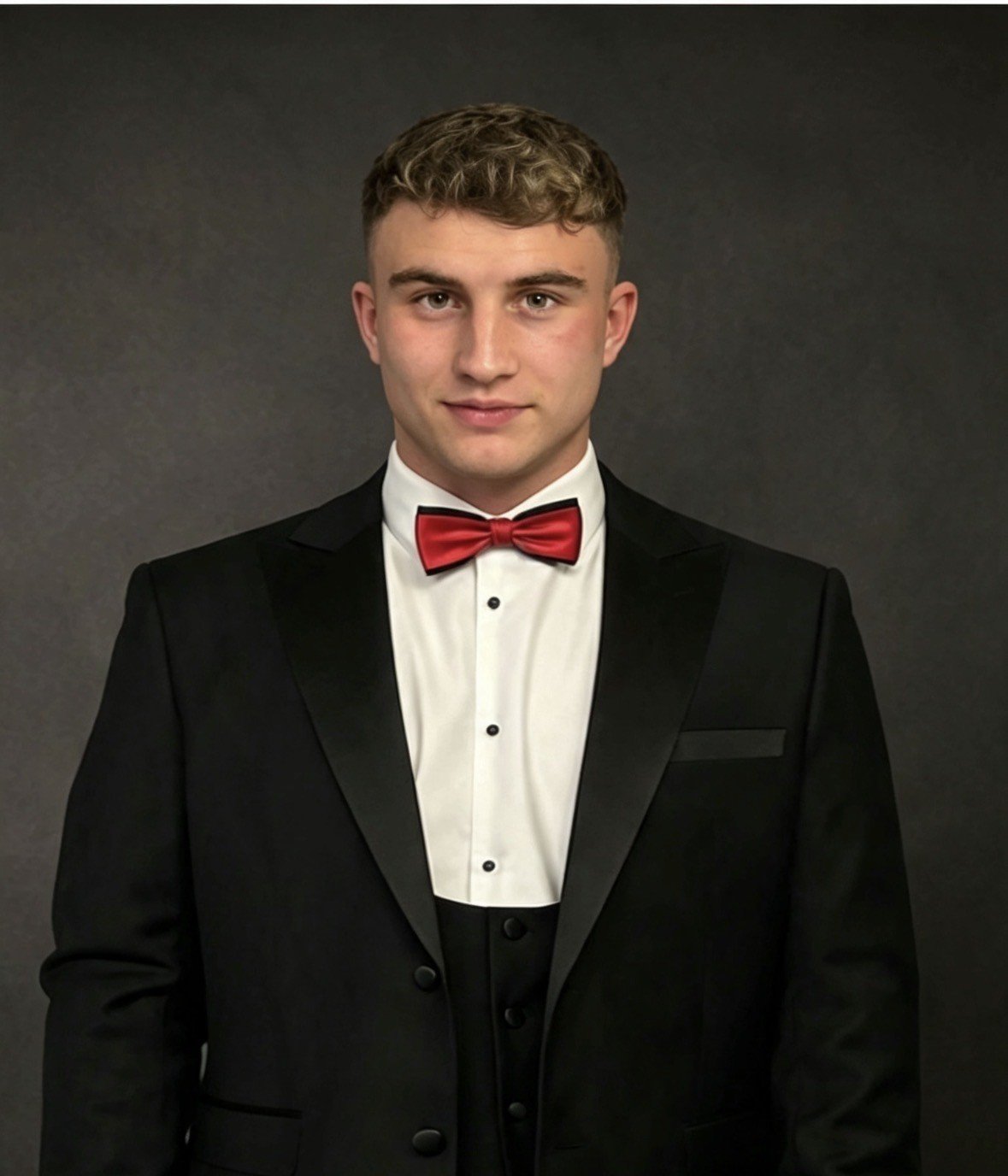 Young man in a black tuxedo with a red bow tie and white dress shirt, standing against a dark background.