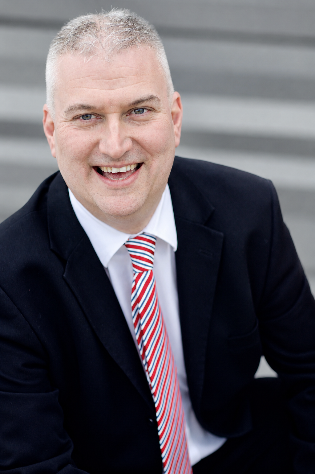 A smiling man wearing a dark suit, white shirt, and red striped tie, sitting outdoors on gray stairs.