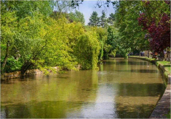 A peaceful river lined with lush green trees and shrubs, with a walking path on the right side and some people in the distance.
