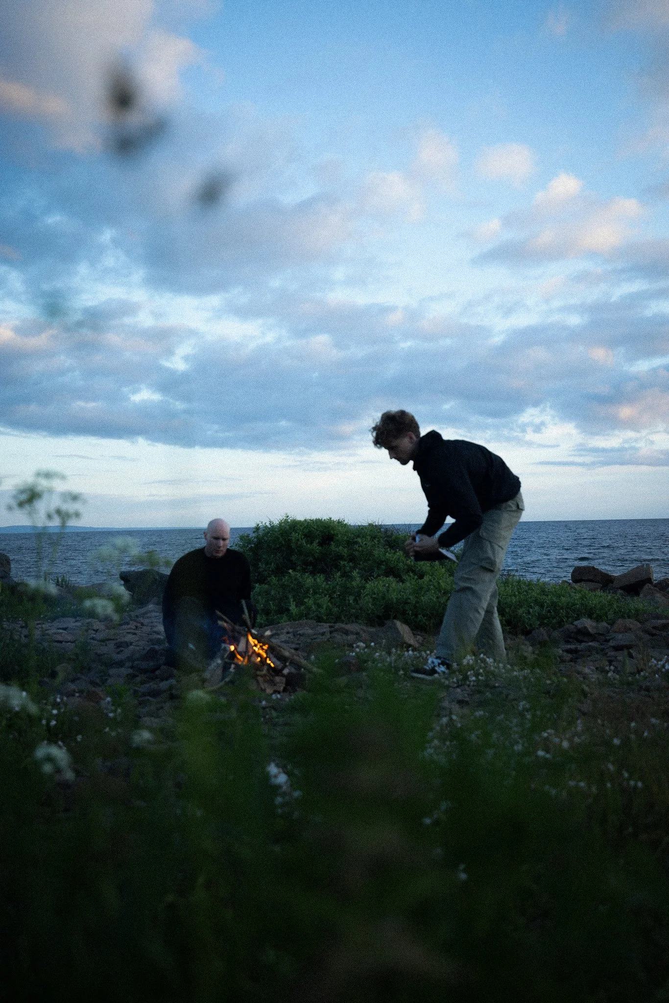 Two men are near a shoreline with a small campfire. One man is wearing a black shirt and sitting on the rocks, while the other is wearing a black jacket and khaki pants, leaning forward. The scene is outdoors with a body of water and cloudy sky in the background.