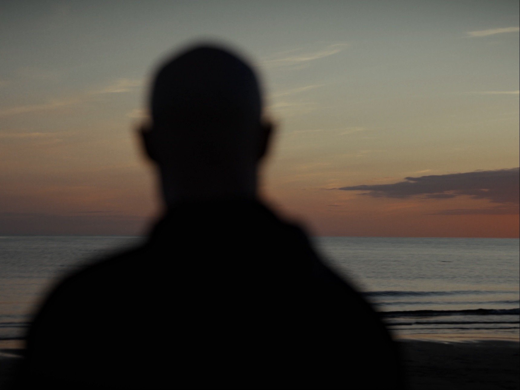 Blurred silhouette of a person facing a sunset over the ocean, with clouds in the sky and a railing in the foreground.