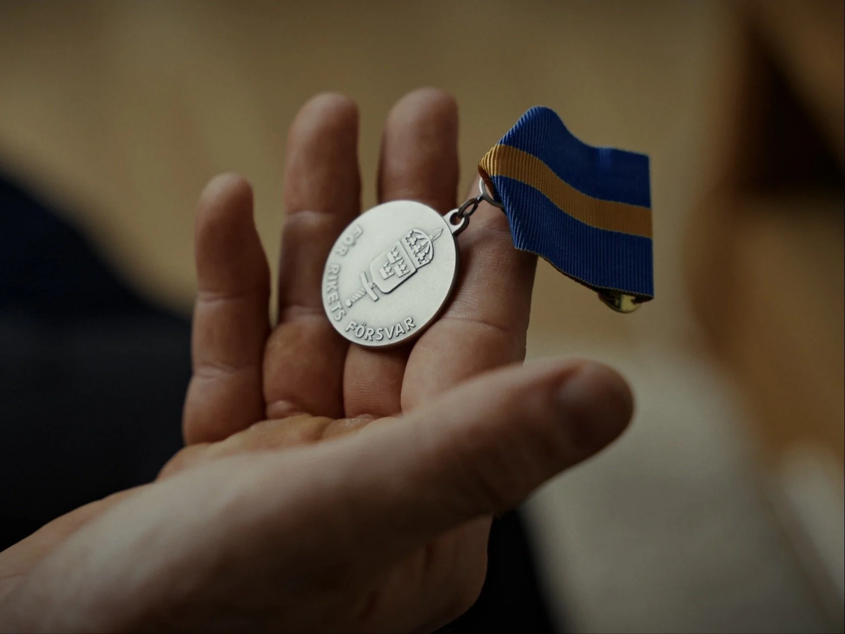 Close-up of a person's hand holding a medal with a blue and yellow ribbon. The medal is silver with an engraved sword and shield and Swedish text that reads 'Försvar'.