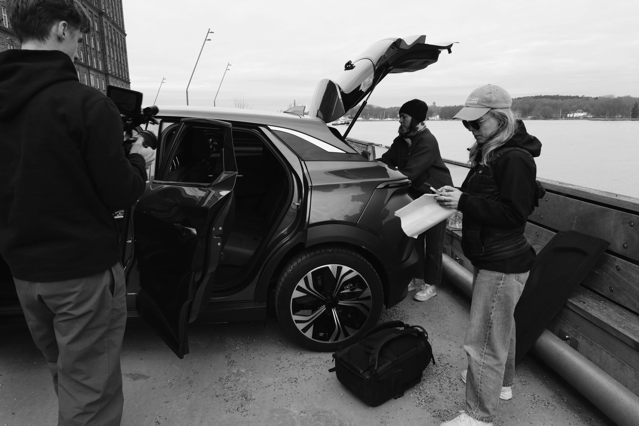 Three people, two women and one man, are standing near an electric vehicle parked by a riverside. The women are looking at tablets, while the man appears to be adjusting a device on the car. The car's trunk is open, and there is a motherboard and other equipment nearby. A bench is next to the river, and there are hills and trees in the background under an overcast sky.