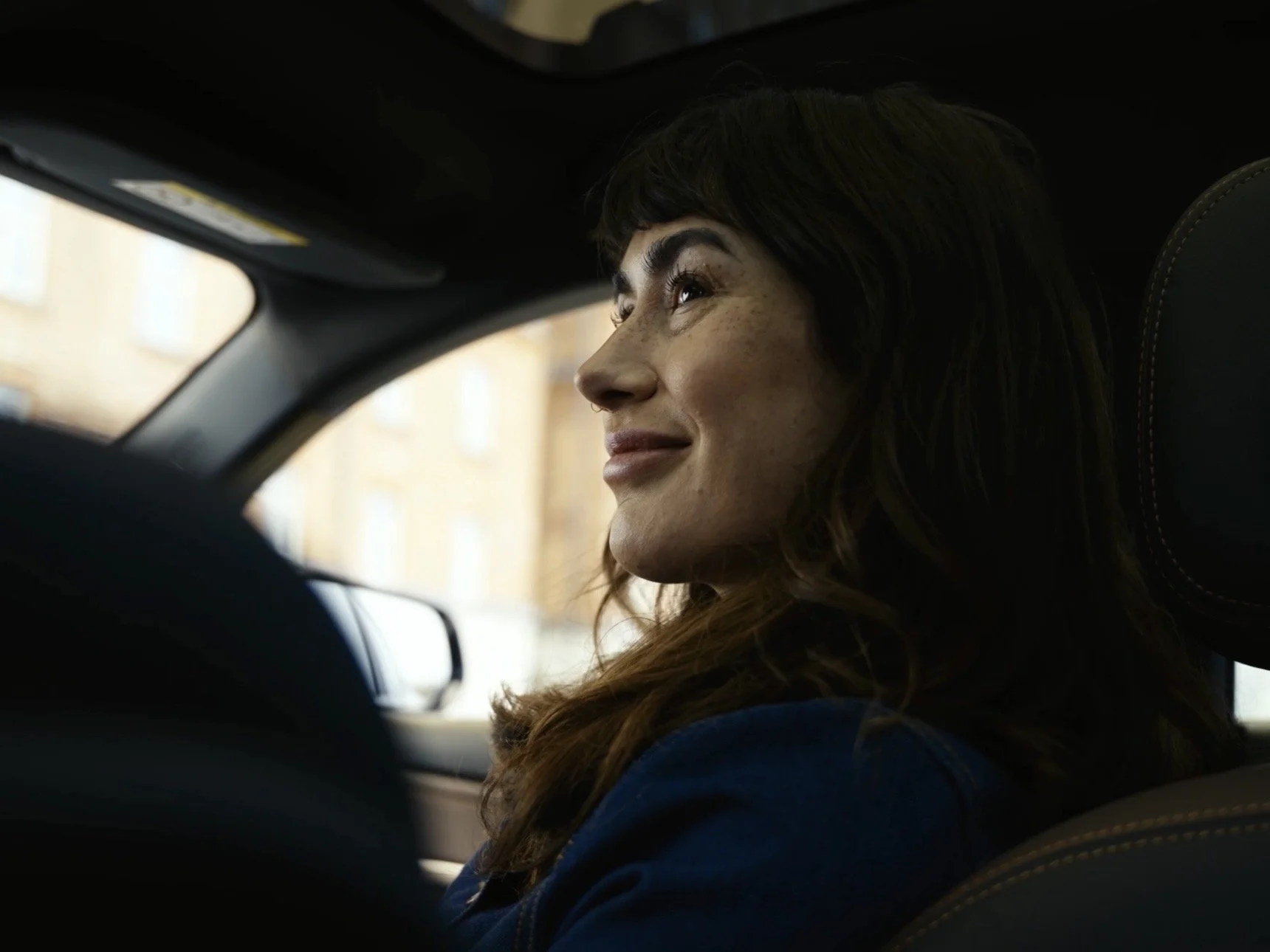 A woman with long brown hair and a subtle smile sitting inside a car, looking out the window.
