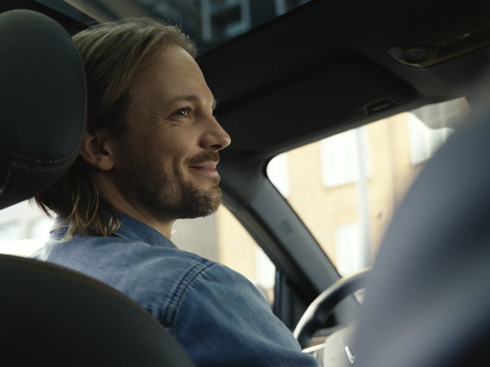 A man with long blonde hair and a beard sitting in the driver's seat of a car, smiling and looking out the window.