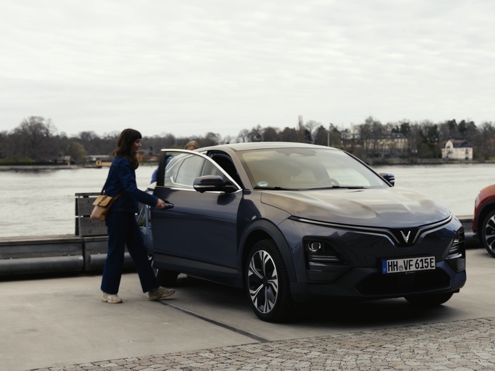 A woman is getting into a dark blue electric vehicle parked on a waterfront promenade with a river and houses in the background.