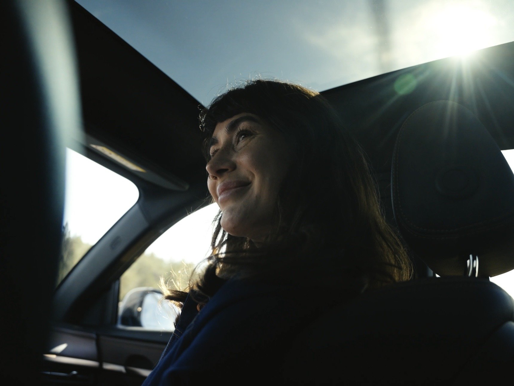 A woman sitting in the passenger seat of a car, smiling and looking upwards with sunlight streaming in from the window above.