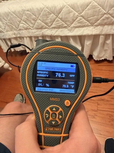 Hand holding a digital meter measuring humidity, temperature, and other environmental data on a room hardwood floor.