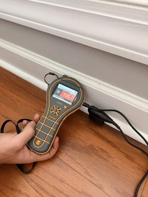 A person holding a digital moisture tester showing a reading of 100.0 meaning fully saturated