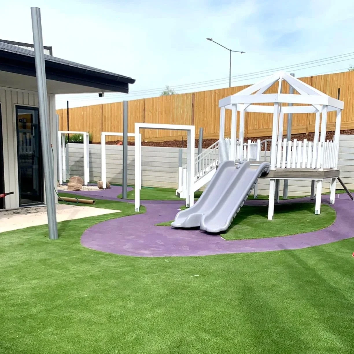 A backyard with artificial grass, a purple pathway, a white playhouse with a slide, a swing set, and a wooden fence.