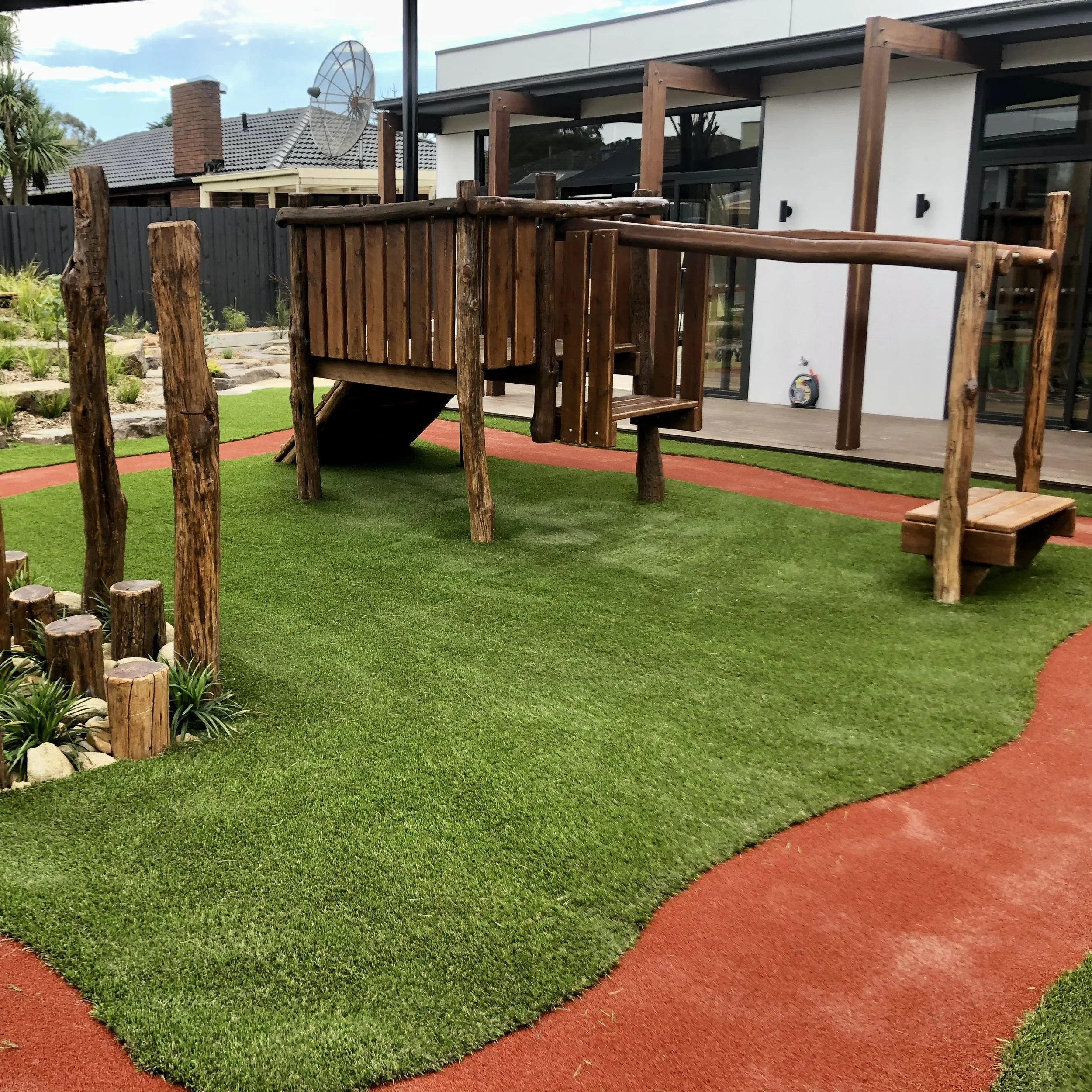 A children's outdoor play area featuring a wooden climbing structure with a slide, surrounded by artificial green grass and a red rubber safety surface. There is a landscaped garden with small plants, and a modern house with large windows and a deck in the background, along with neighboring rooftops and a satellite dish.