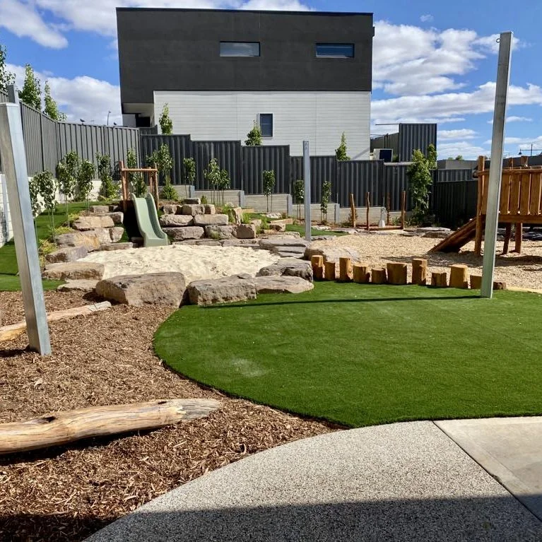 Children's playground with a small slide, rocks, artificial turf, sand area, and wooden climbing structures in a backyard. Modern house with black and white exterior and trees in the background.