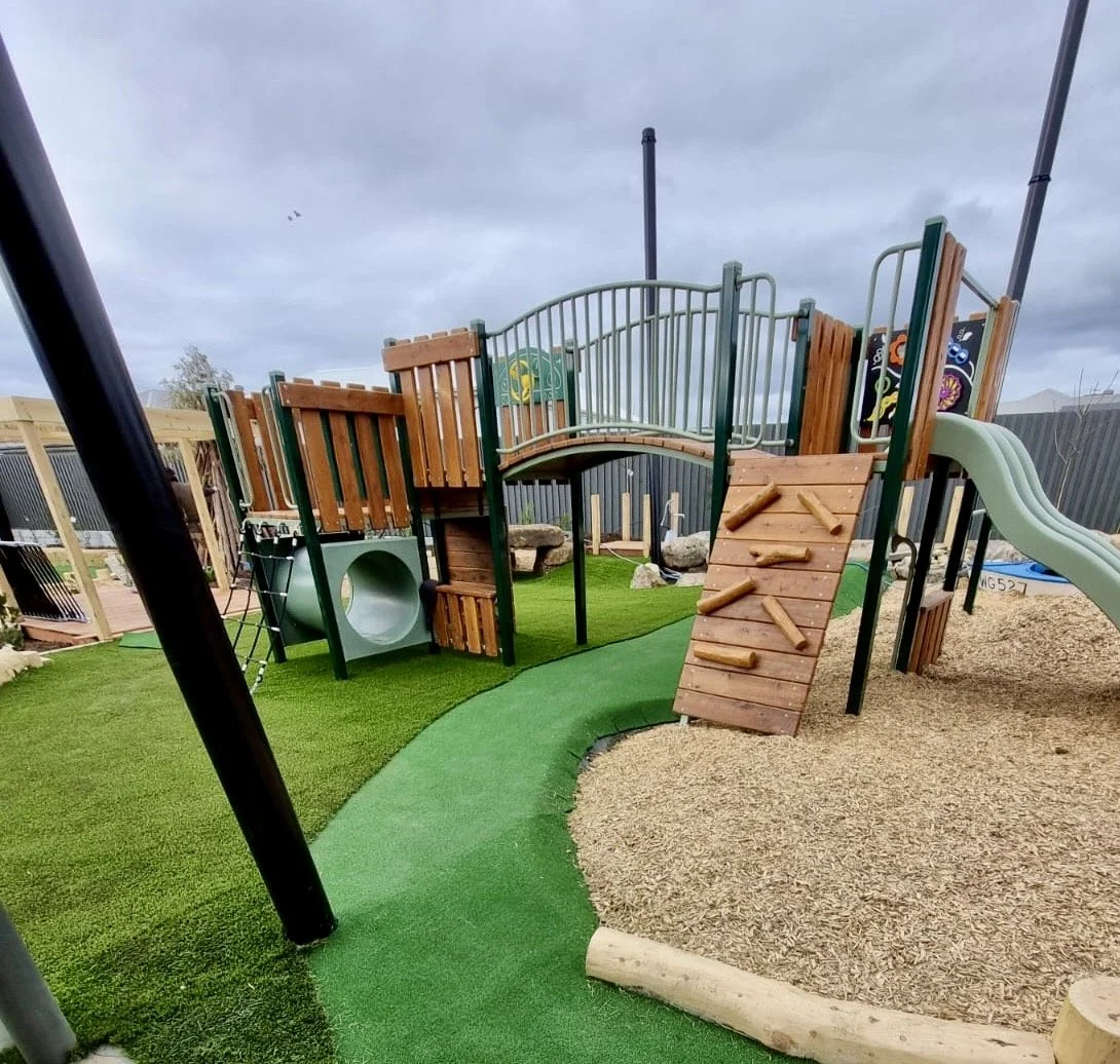 Children's playground with wooden and metal structures, green slide, climbing wall, artificial grass, and wood chips on the ground.