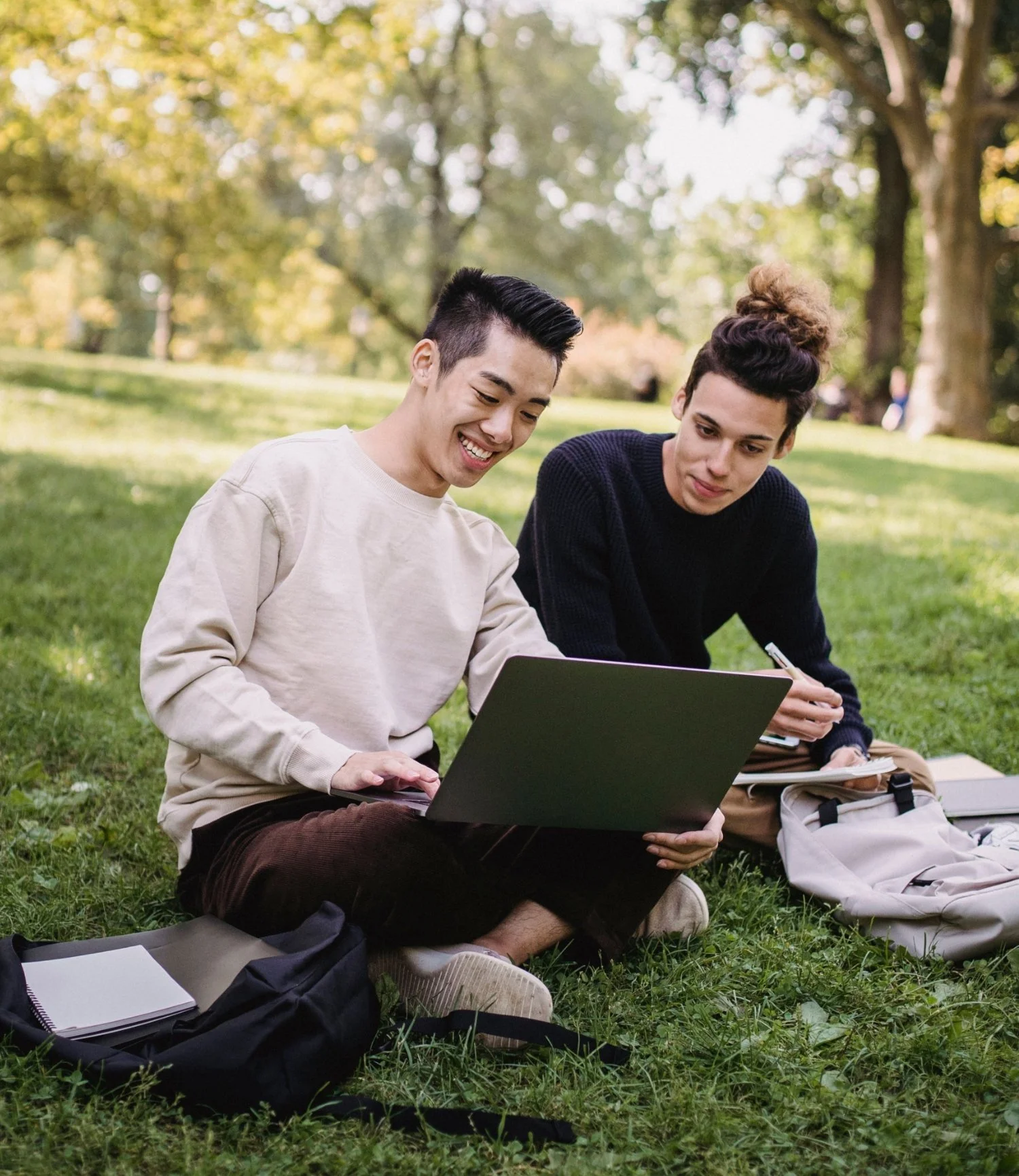 Two men sitting on grass at a college campus, looking at a laptop screen and smiling.
