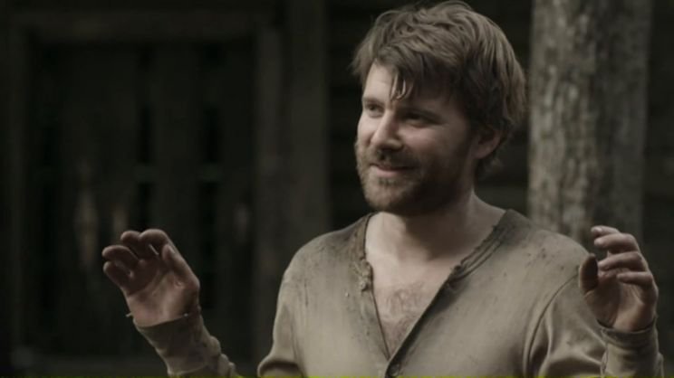 A man with a beard and disheveled hair, wearing a dirty beige shirt, smiling and gesturing with his hands outdoors near trees.