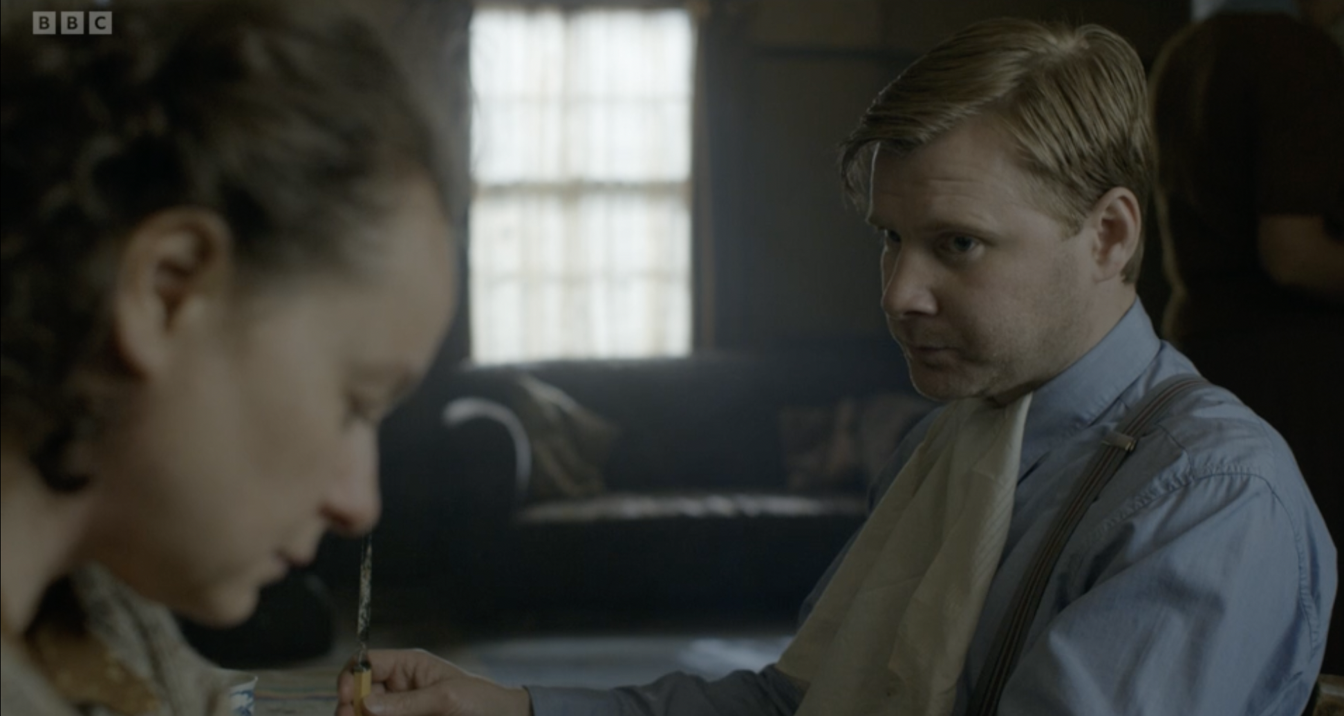 A young man and woman sit at a table in a dimly lit room. The woman with long wavy hair is in the foreground, and the man with short hair and a serious expression is facing her. The woman appears to be preparing to have an injection or shot, with the man's hand holding her arm.