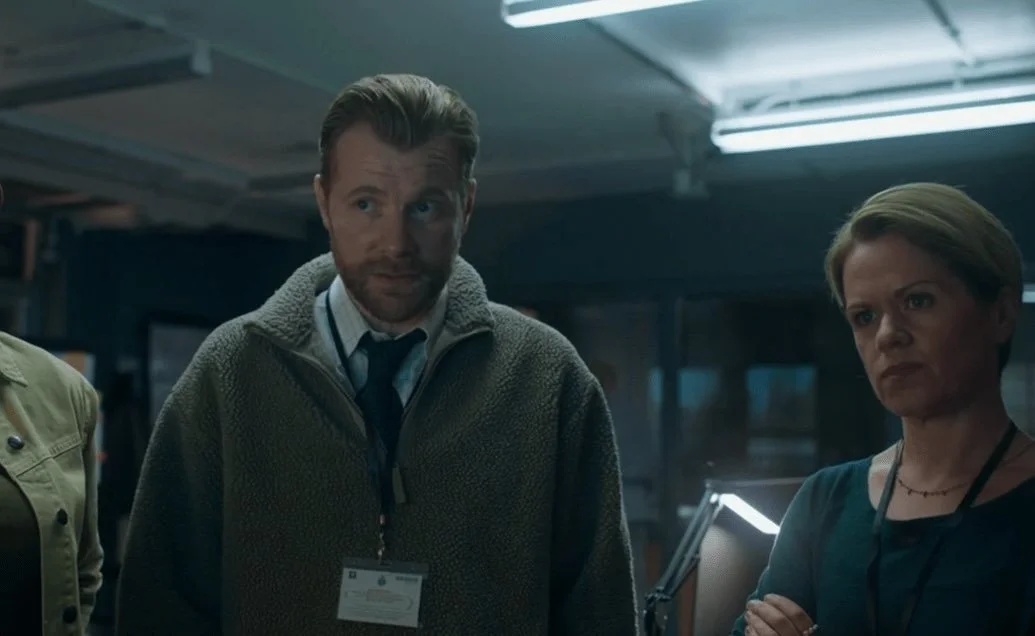 Three people, two women and one man, standing indoors in a dimly lit room, possibly an office or control center, with serious expressions.