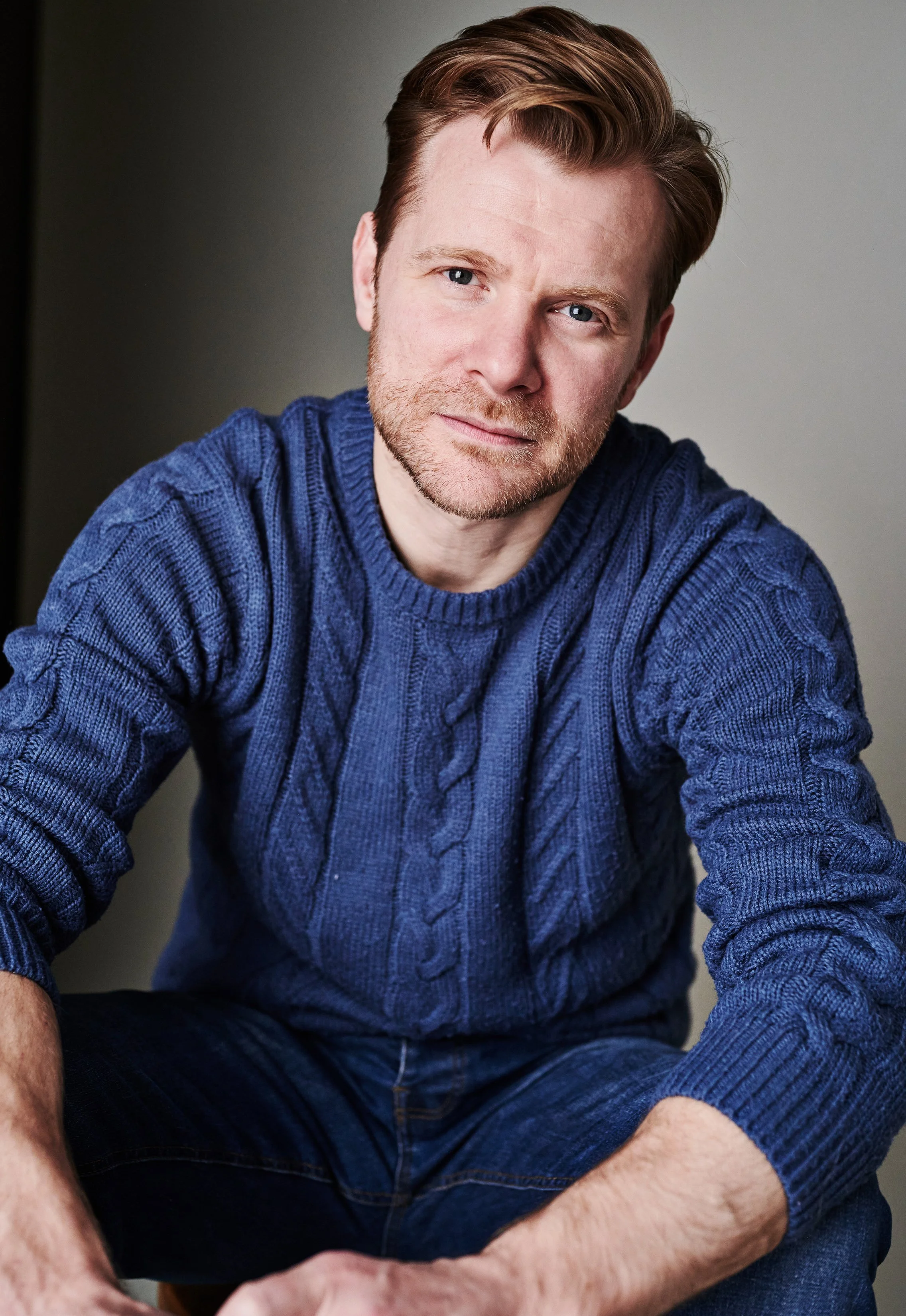 A man with light skin, reddish-brown hair, and a beard wearing a blue knitted sweater and dark shorts, sitting and looking at the camera.