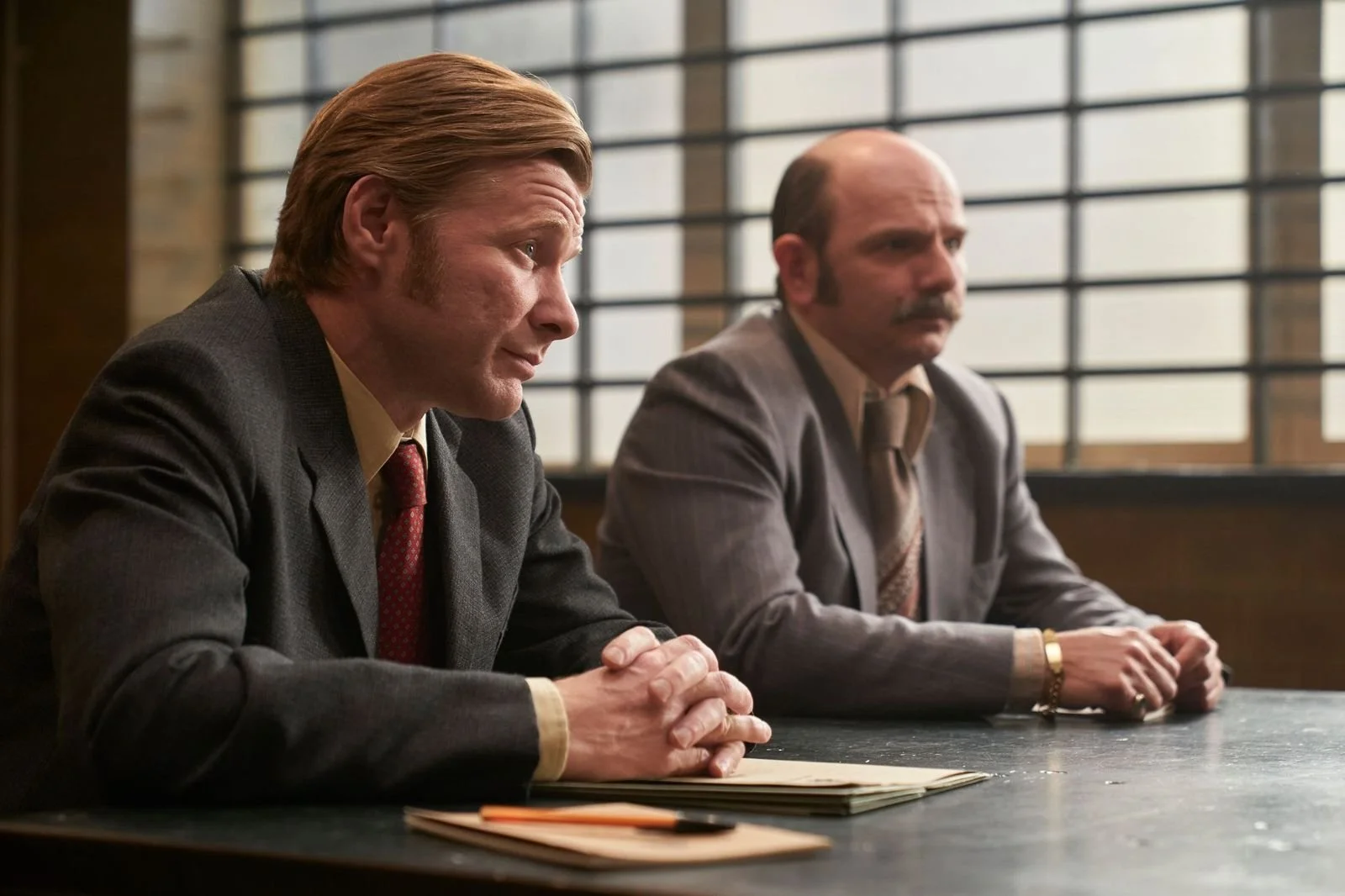 Two men in suits sit at a table in a room with bar window behind them, appearing to be in a formal discussion or interview.