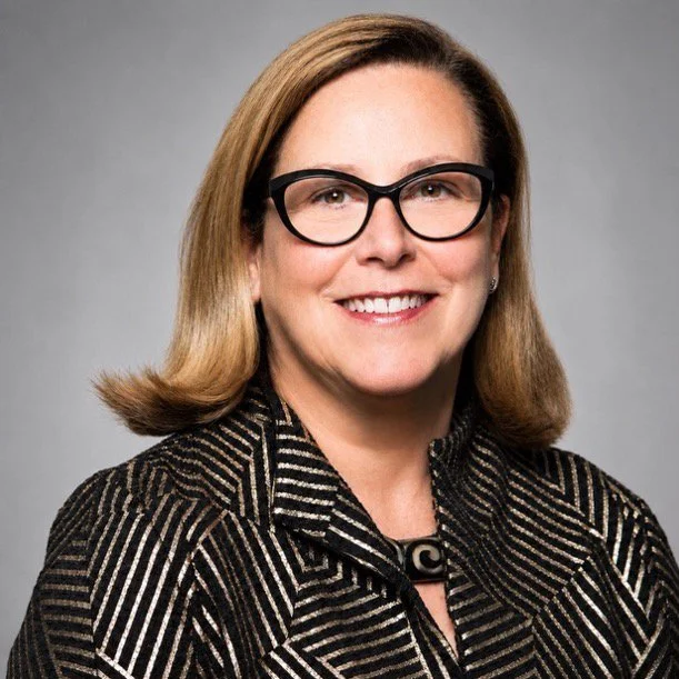 Professional woman with shoulder-length blonde hair, black glasses, and a black and white patterned blazer, smiling against a gray background.