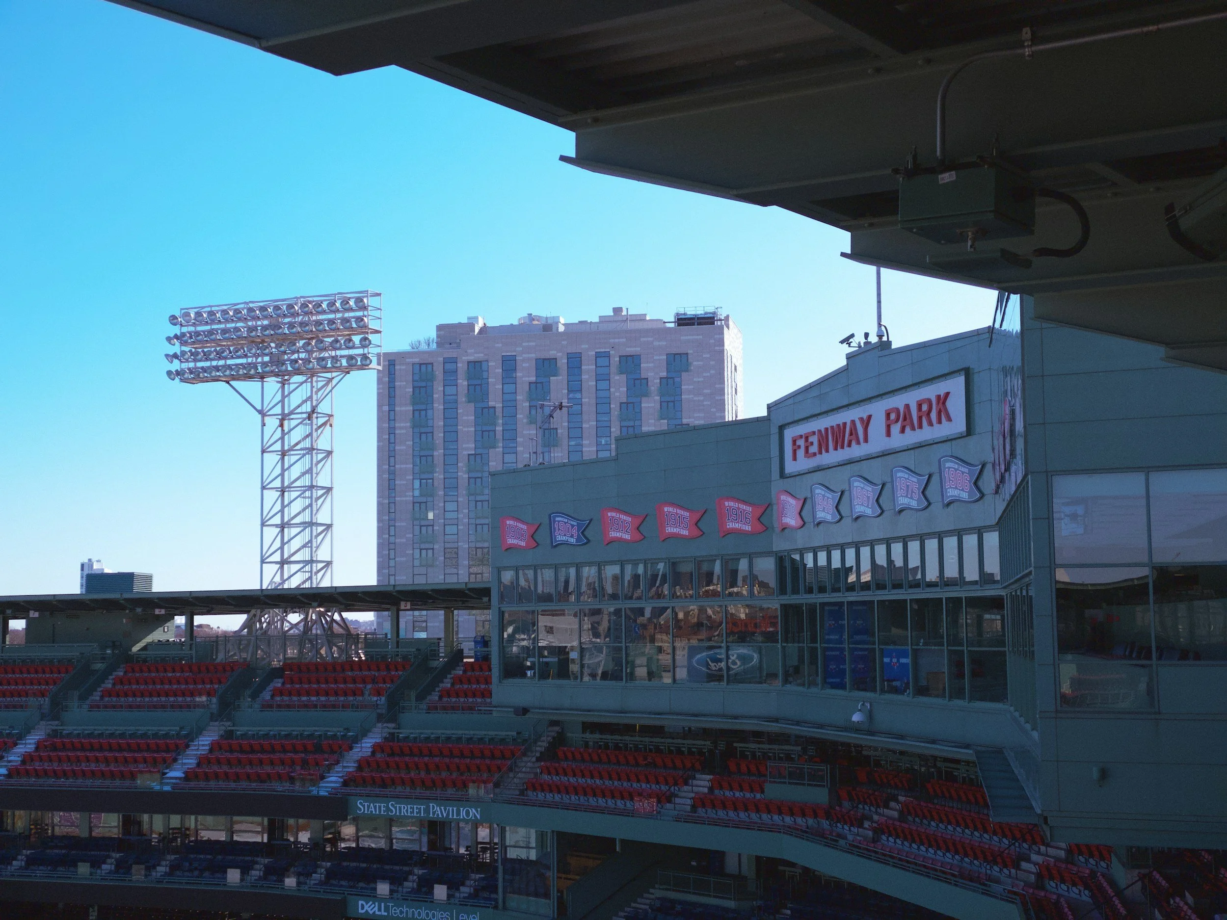 View of Oracle Park stadium with red seats in the stands, a billboard with the wording 'Fenway Park', floodlights, and a backdrop of tall buildings under a clear blue sky.