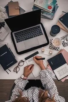 Top view of a person writing in a notebook surrounded by a laptop, books, glasses, a coffee mug, and stationery on a cluttered desk.