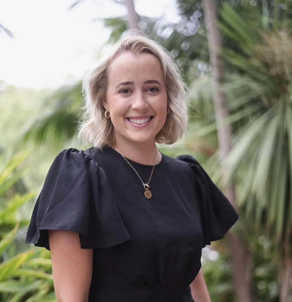 A woman with shoulder-length blonde hair, wearing a black dress with puffed sleeves, smiling outdoors with green tropical plants in the background.