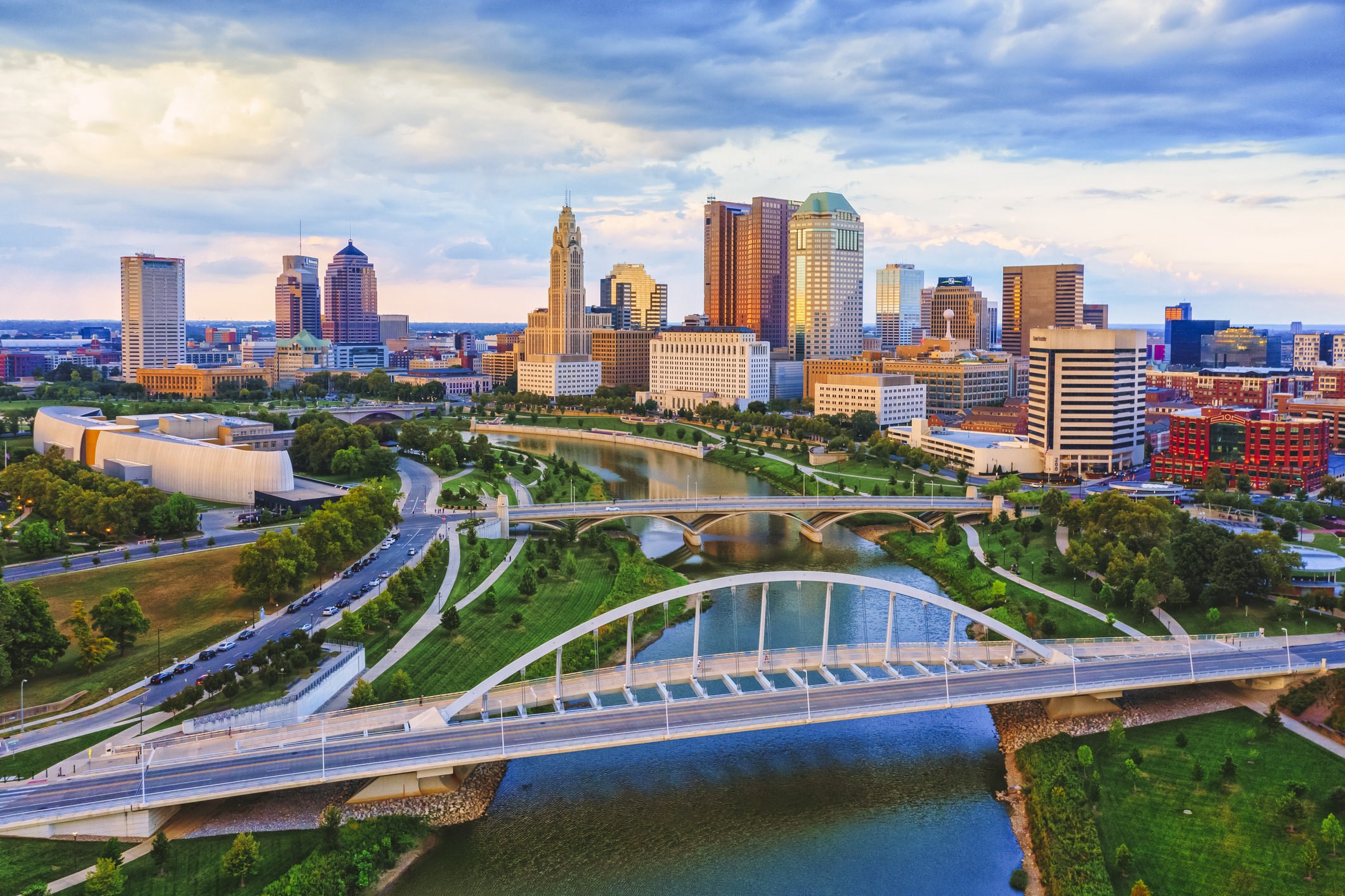 A city skyline with tall buildings, a river, and bridges, under a partly cloudy sky during the daytime.