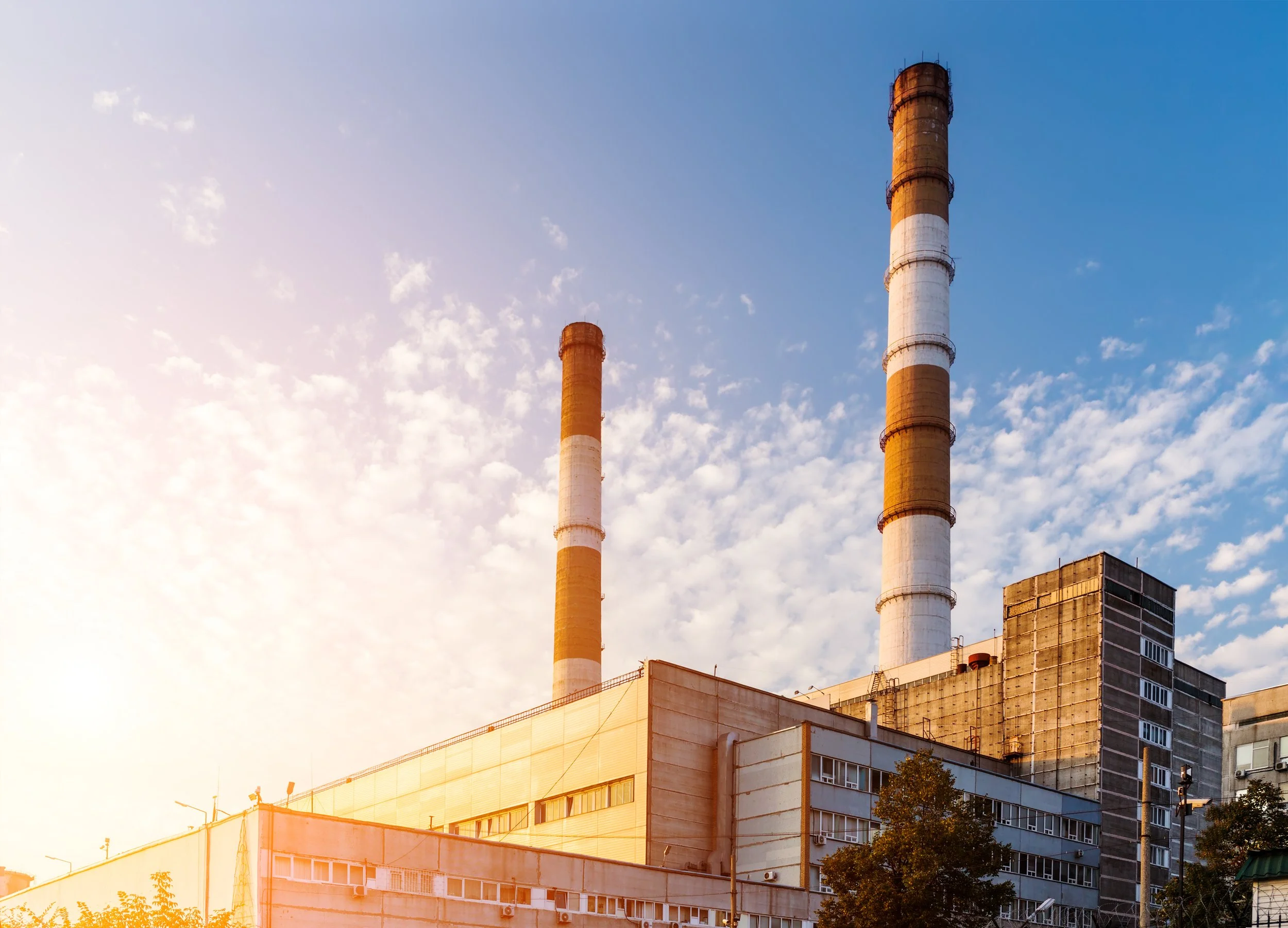 Image of industrial buildings with tall chimneys against a partly cloudy sky during the day.