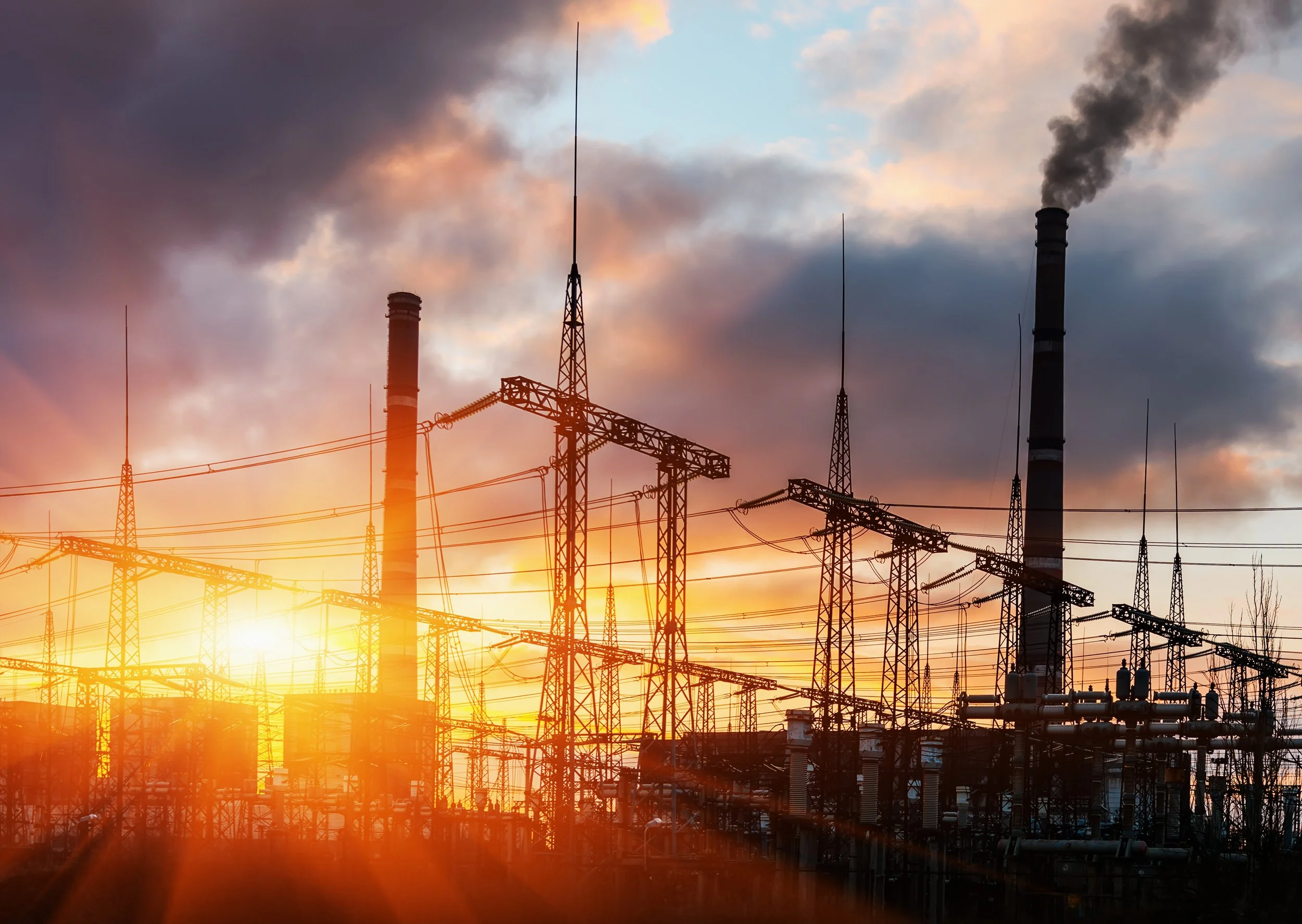 Industrial power plant with multiple smokestacks emitting dark smoke during sunset, with a complex network of electrical transmission towers and wires in the foreground.