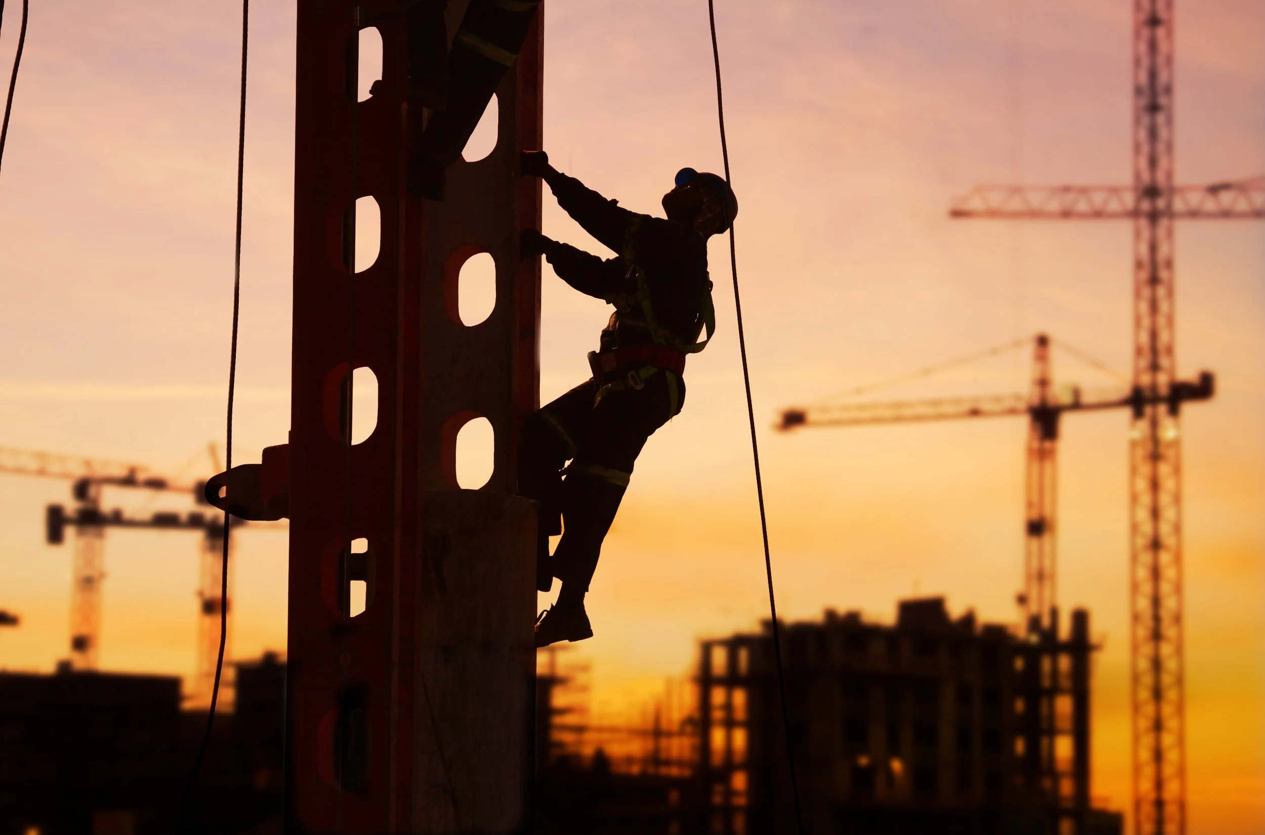 Silhouette of a construction worker climbing on a utility pole during sunset, with cranes in the background.