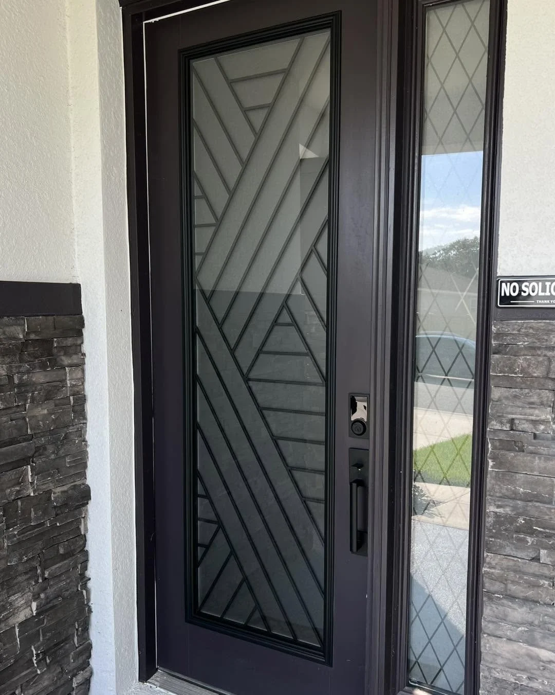 Front door with decorative glass and geometric black metal design, flanked by stone and stucco walls, with a security keypad and a 'No Soliciting' sign.