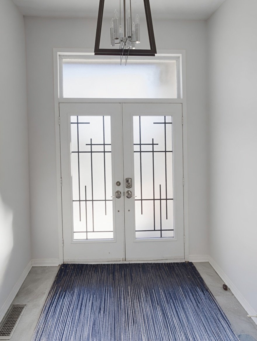 White front door with decorative black metal grille, frosted glass window above, located in a small entryway with white walls and a striped dark blue and gray rug on a concrete floor.