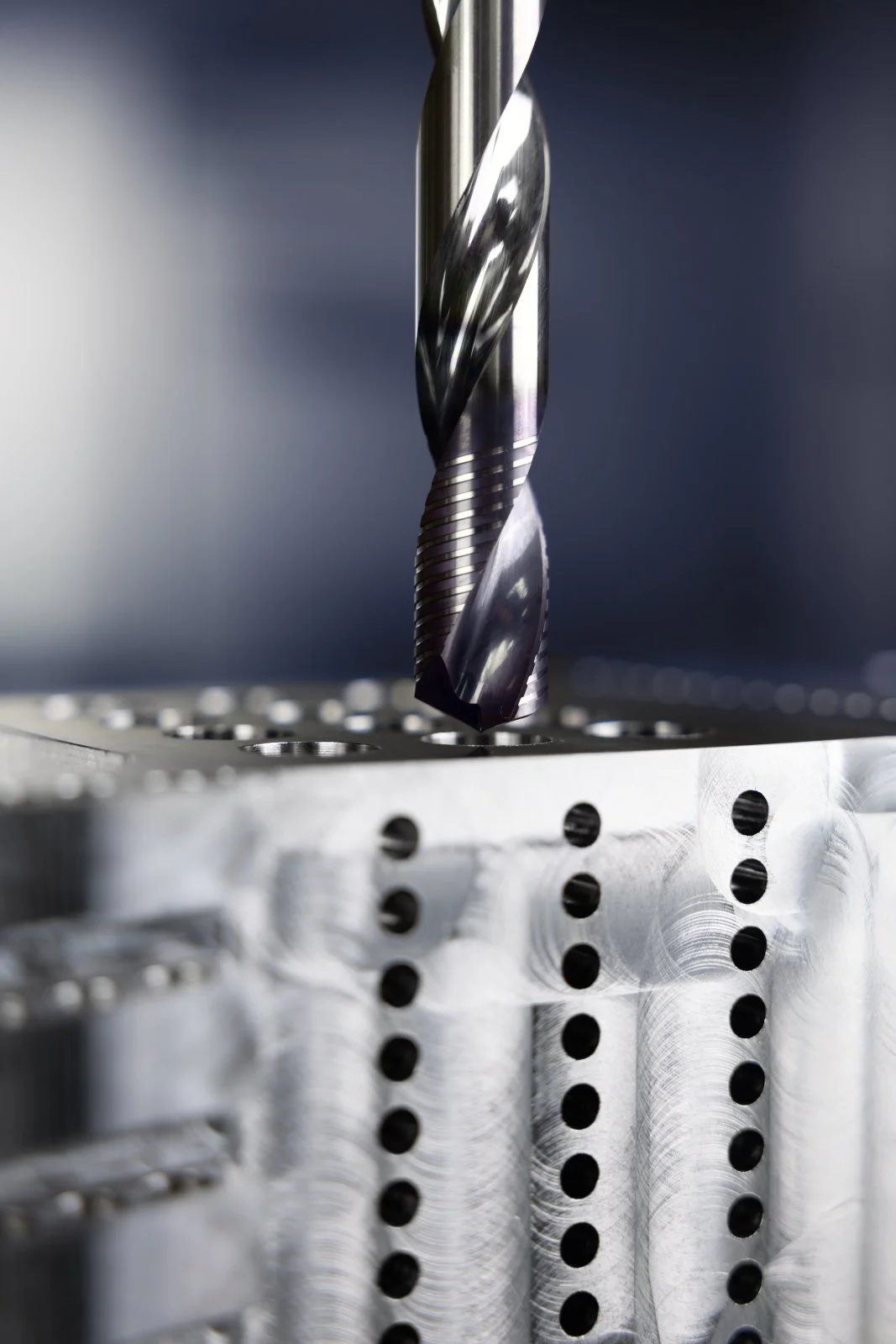 Close-up of a titanium drill bit above a metallic work surface with drilled holes.