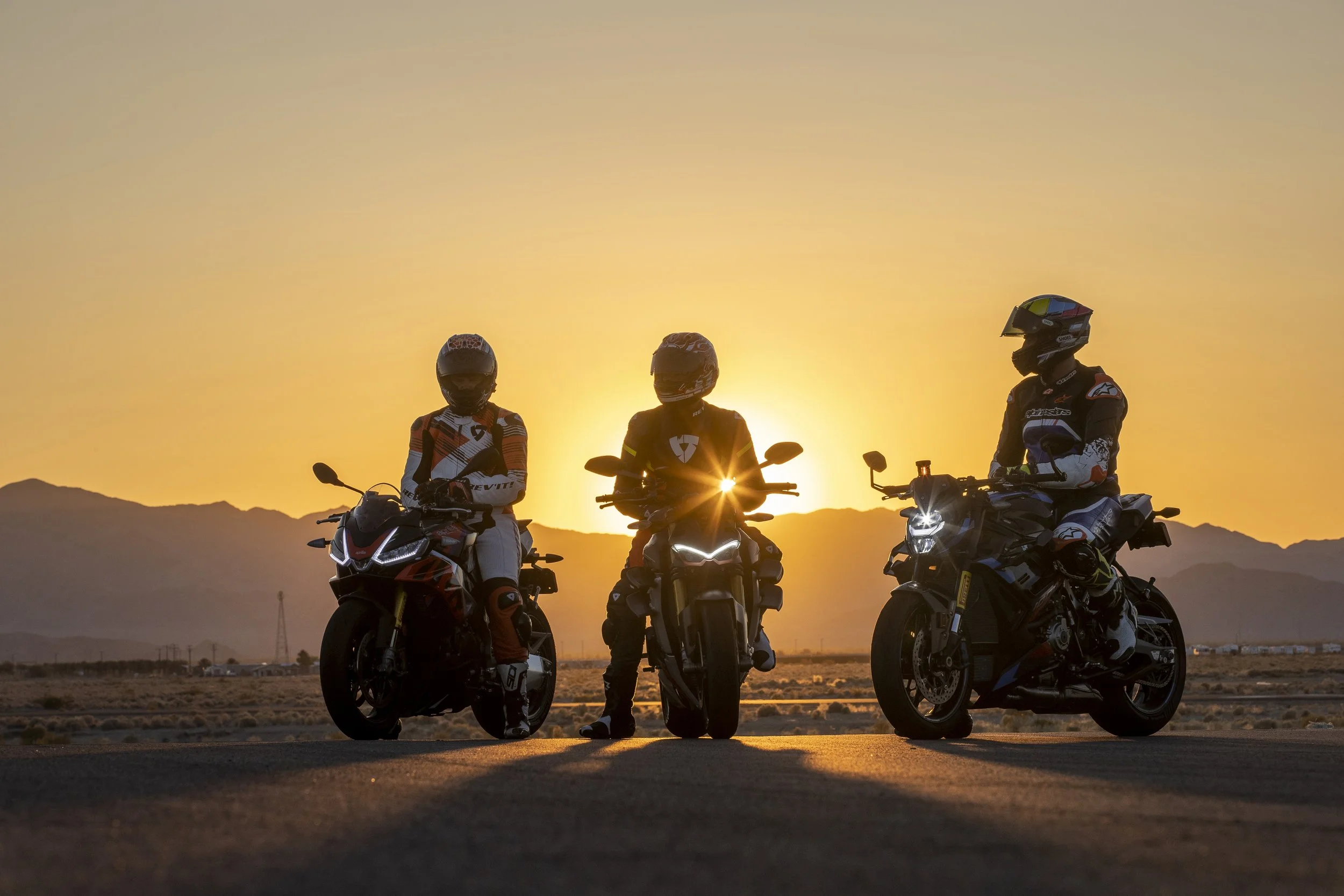 Three motorcyclists wearing helmets and riding gear sitting on their motorcycles at sunset with mountains in the background.