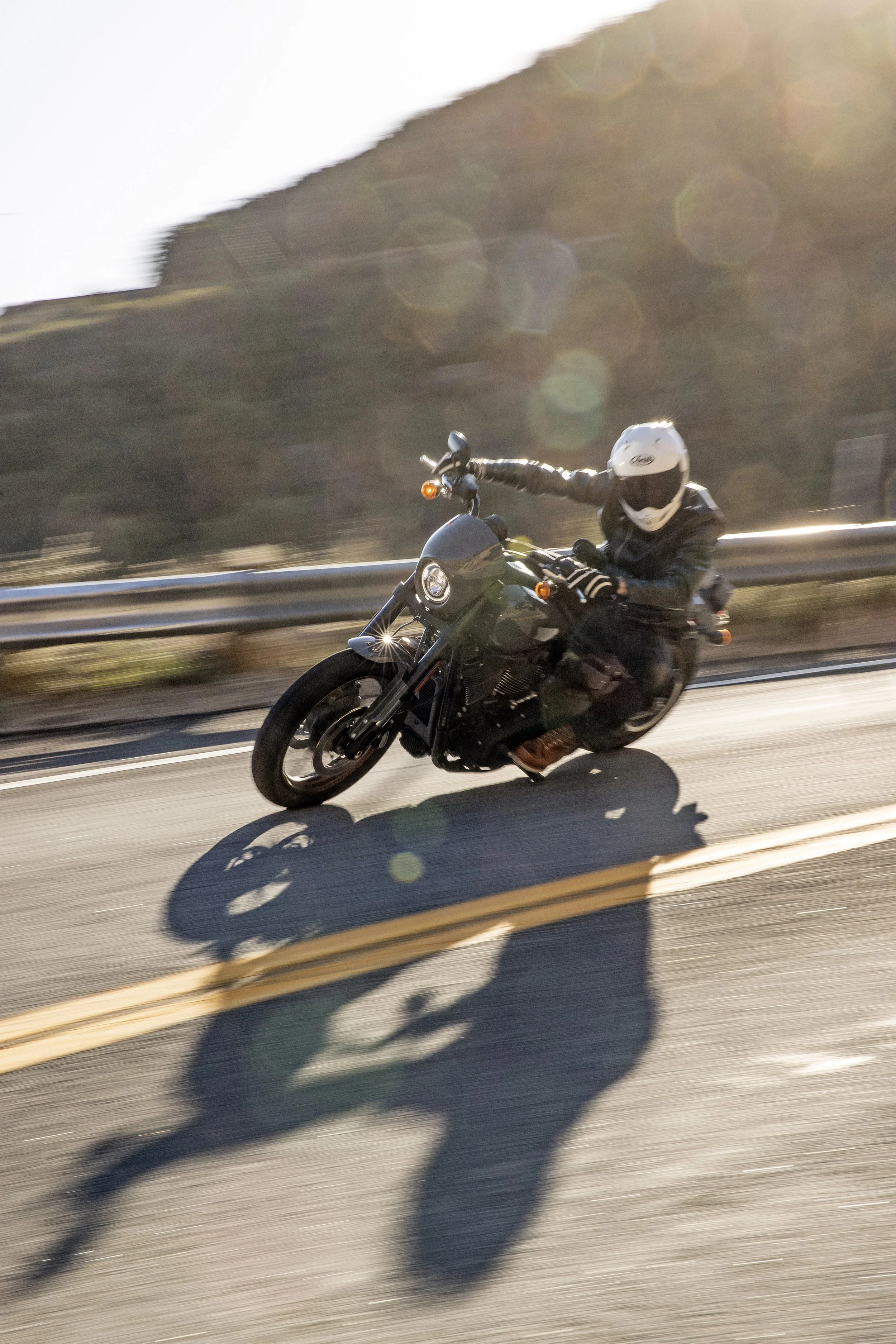 Motorcyclist wearing a helmet and black leather gear riding on the road with a shadow cast on the pavement, blurred background of hills and guardrail.