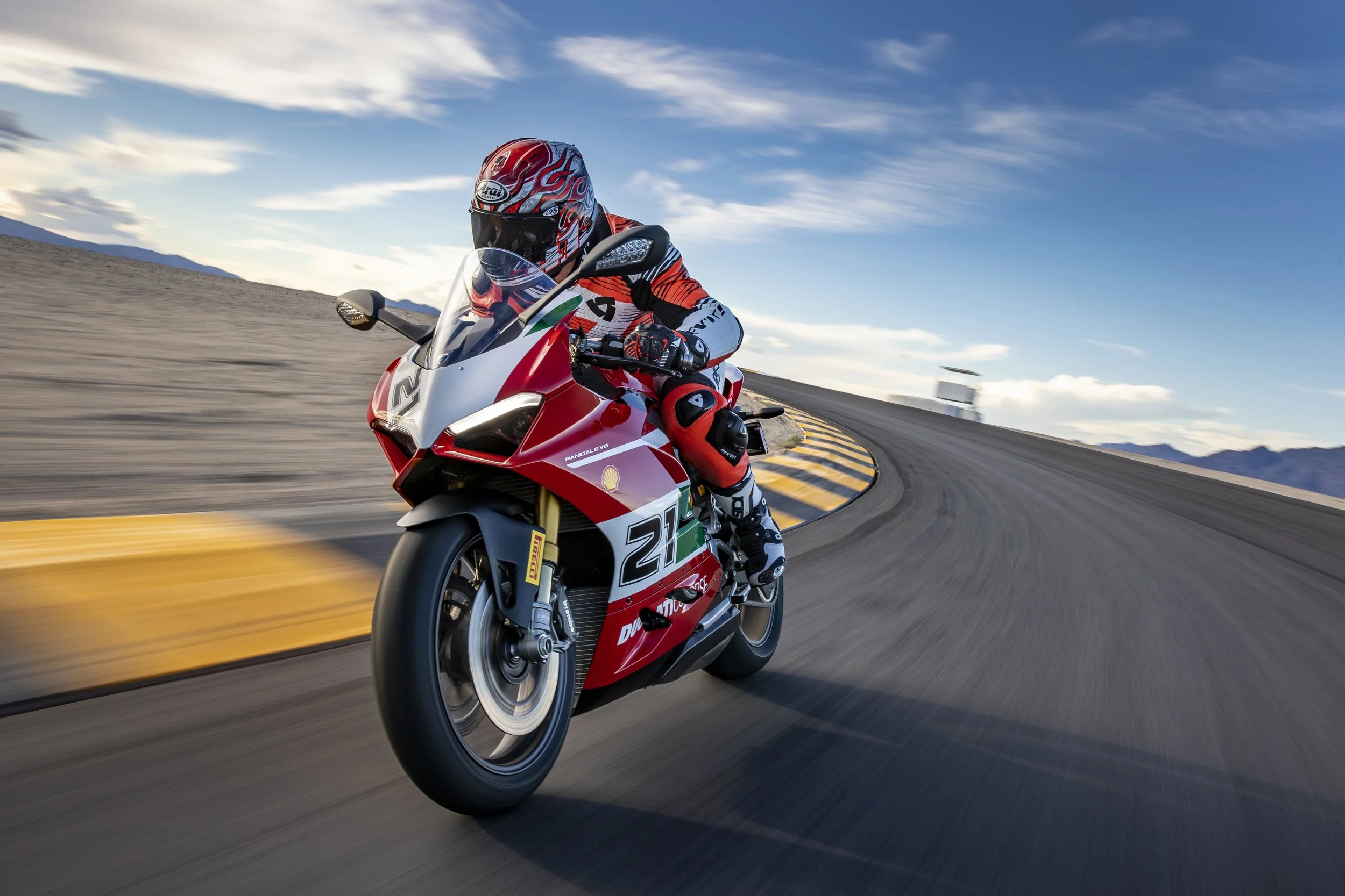 Motorcycle racer in full gear riding a red and white racing motorcycle on a curving road with mountains in the background and a partly cloudy blue sky.