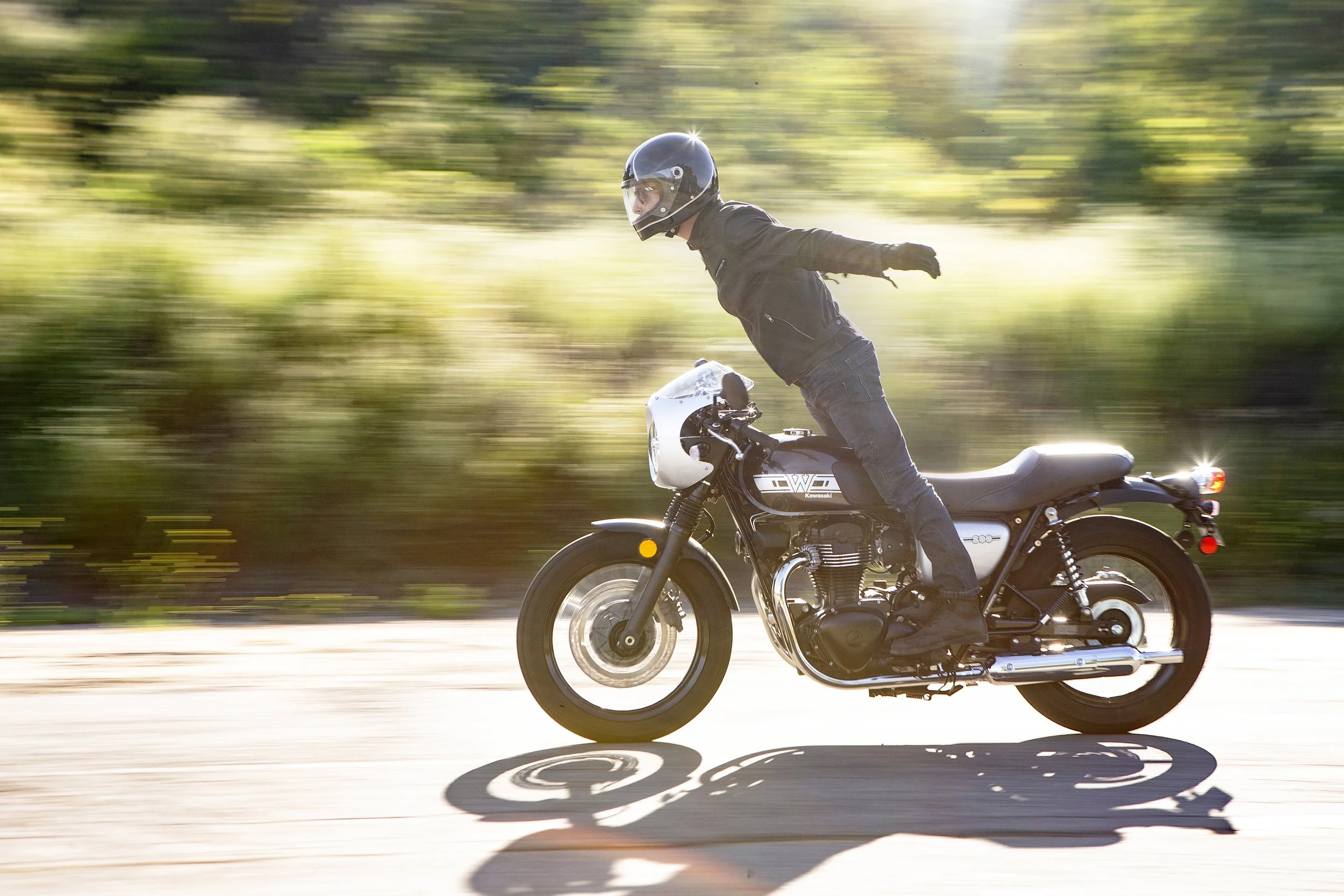 A person wearing a helmet and black clothing is riding a black motorcycle on a road with blurred green trees and sunlight in the background.