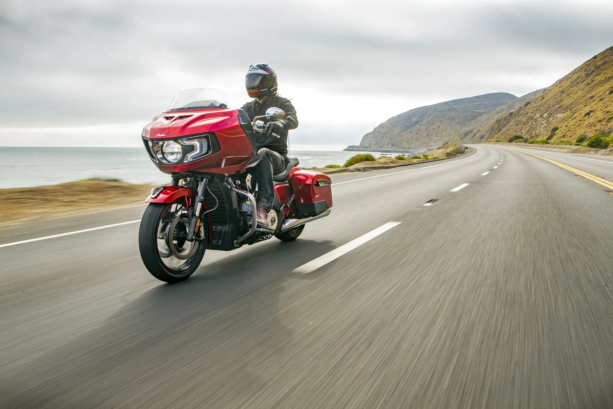 A motorcyclist wearing a black helmet and jacket riding a red touring motorcycle along a coastal highway with cliffs and ocean in the background.