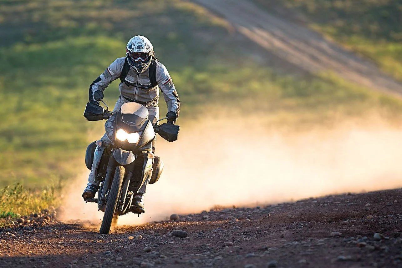 A motorcyclist riding on a dirt trail with dust and grass in a rural setting, wearing a helmet, goggles, and protective gear.