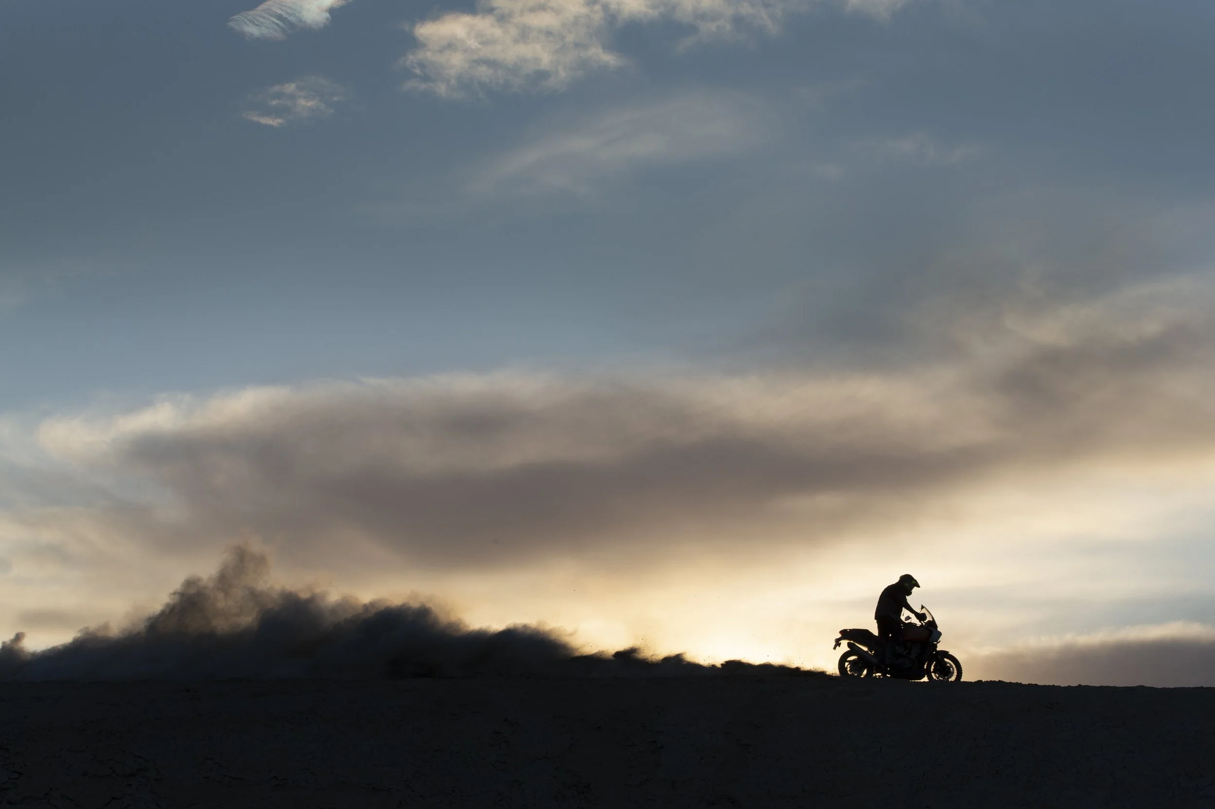 A silhouette of a person riding a motorcycle during sunset or sunrise with a sky filled with clouds and smoke or dust in the background.