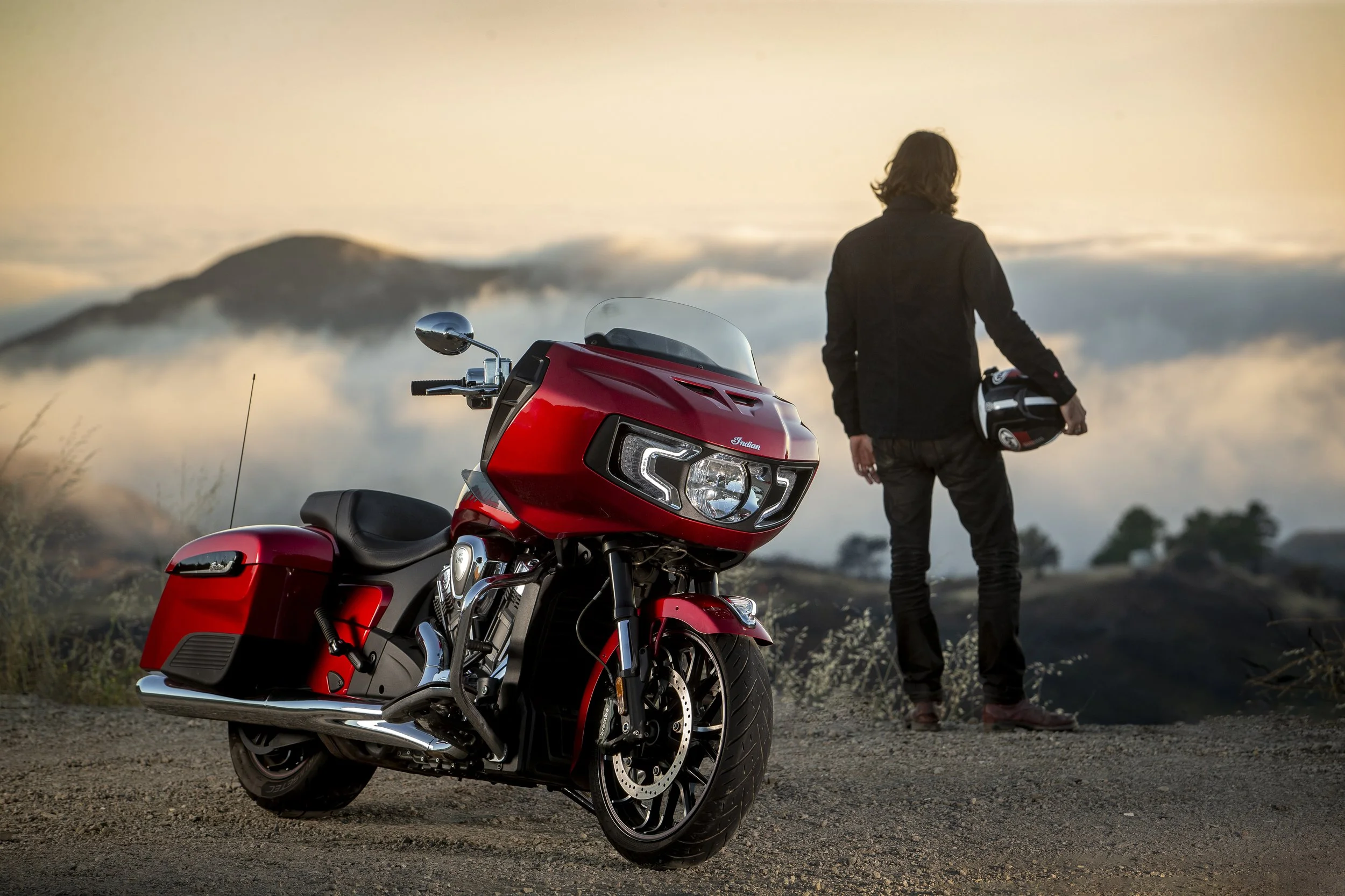 A person with shoulder-length hair, wearing a black jacket, holding a helmet, standing next to a red Indian motorcycle on a dirt road, with a scenic mountain and cloudy sky in the background during sunset or sunrise.